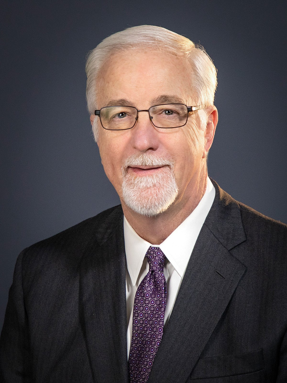 Professional portrait of an older man with white hair and glasses in a dark suit.