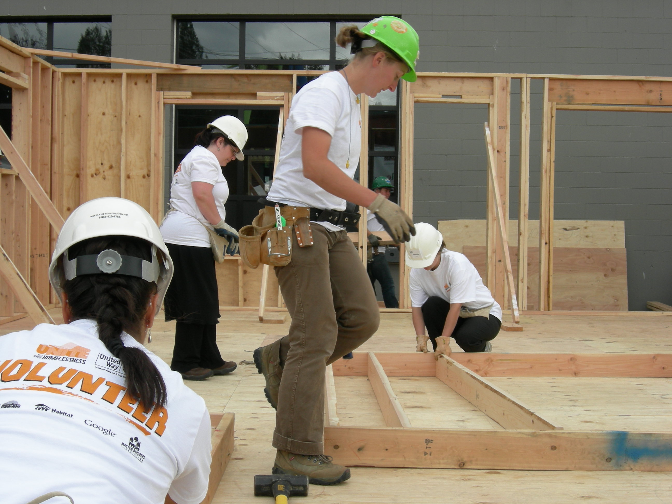 Several volunteers in hard hats and white volunteer t-shirts working on a wooden house frame.