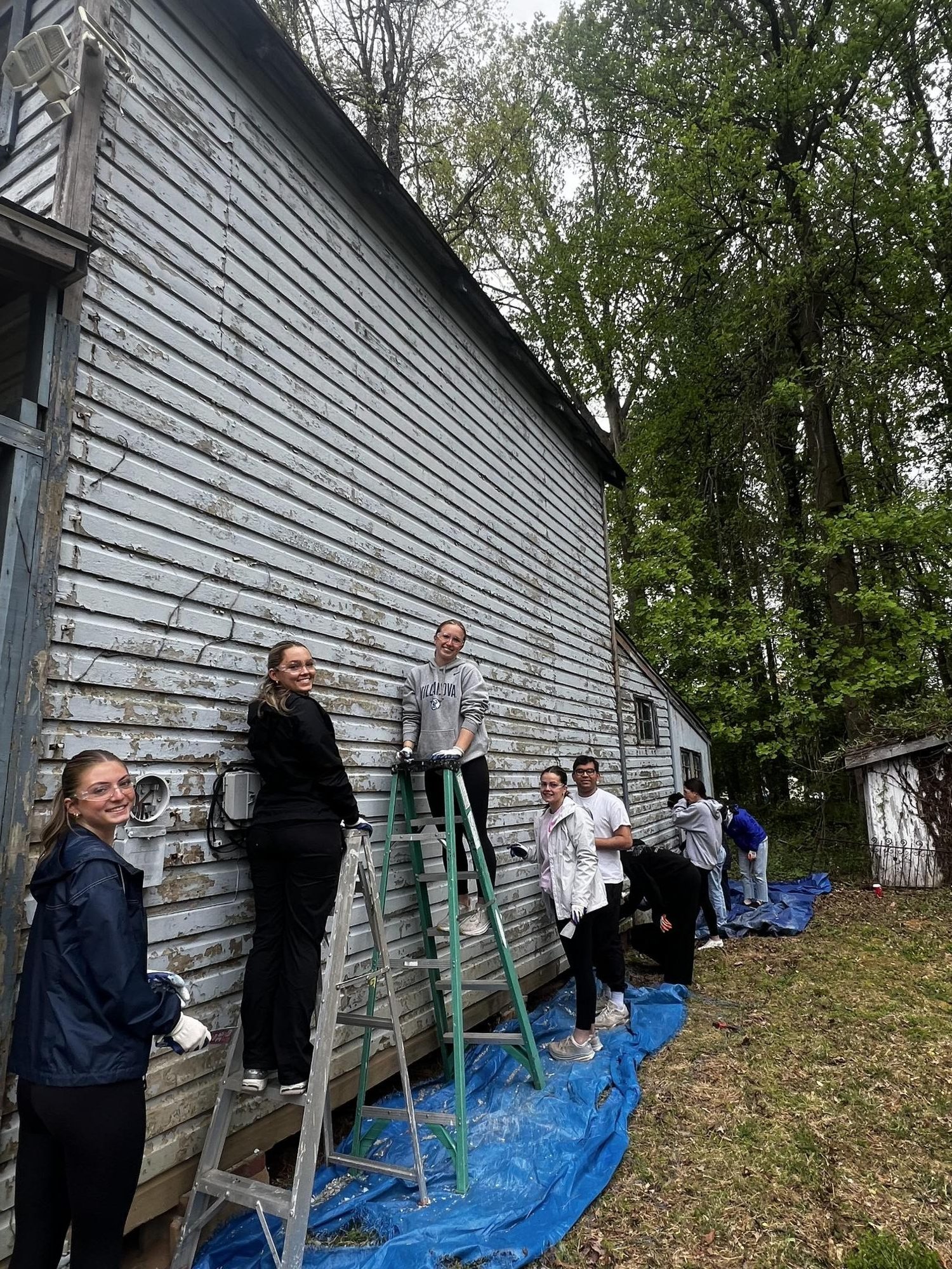 Volunteers on a ladder and on the ground scraping peeling paint from a building's exterior siding.