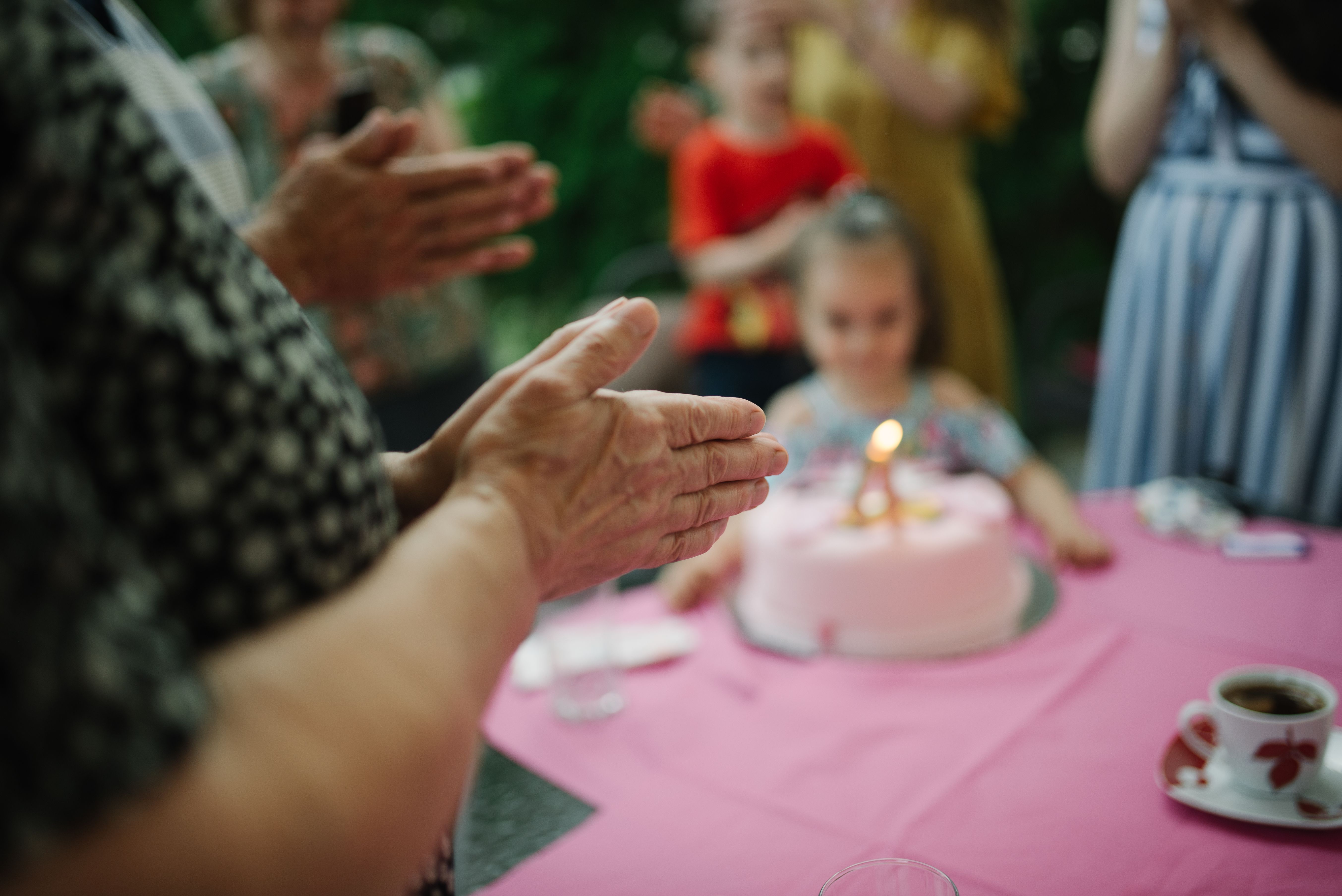 People clapping around a pink birthday cake with a lit candle outdoors.