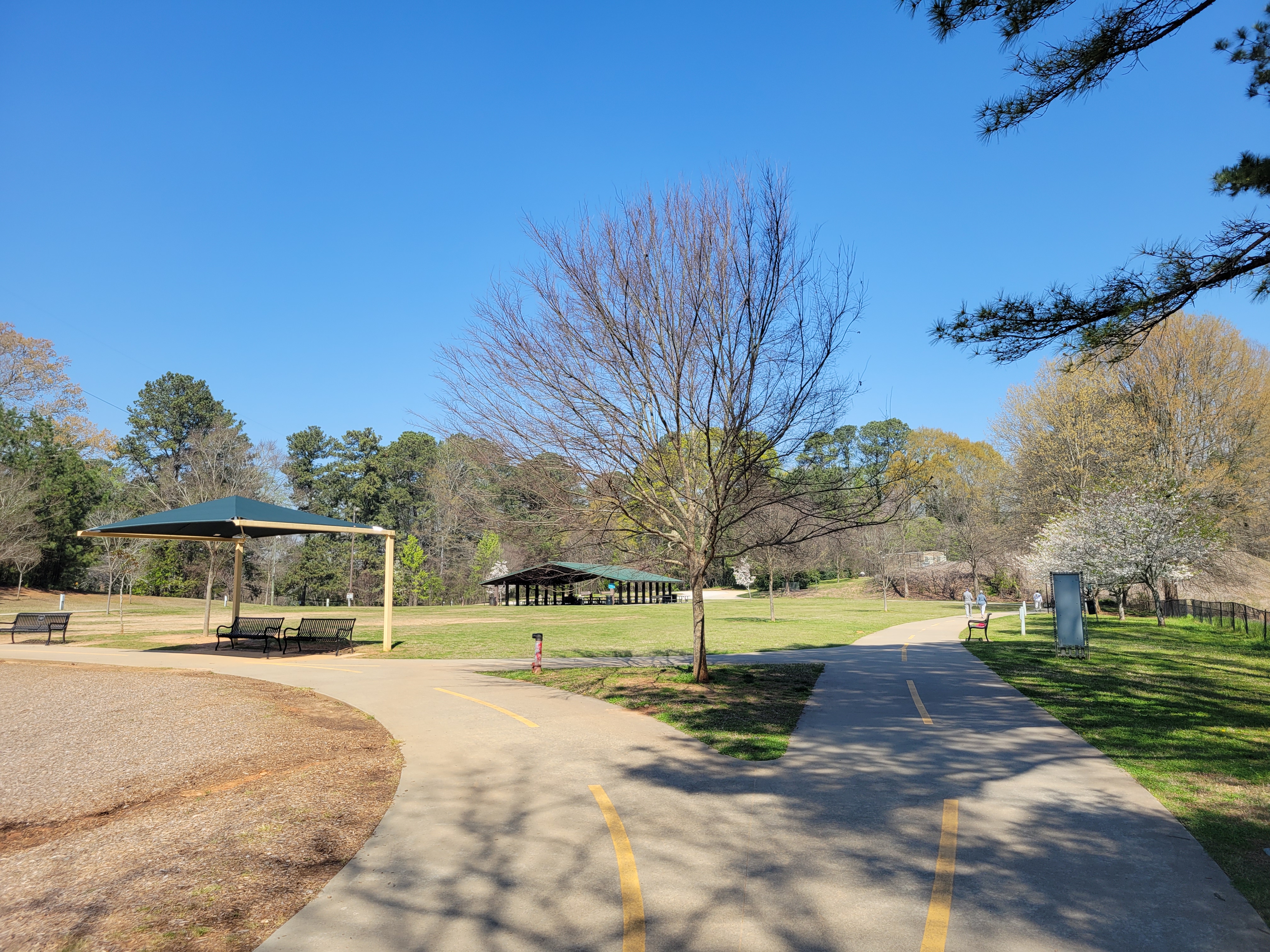 Paved walking path through a park with pavilions and open grassy areas under blue sky.