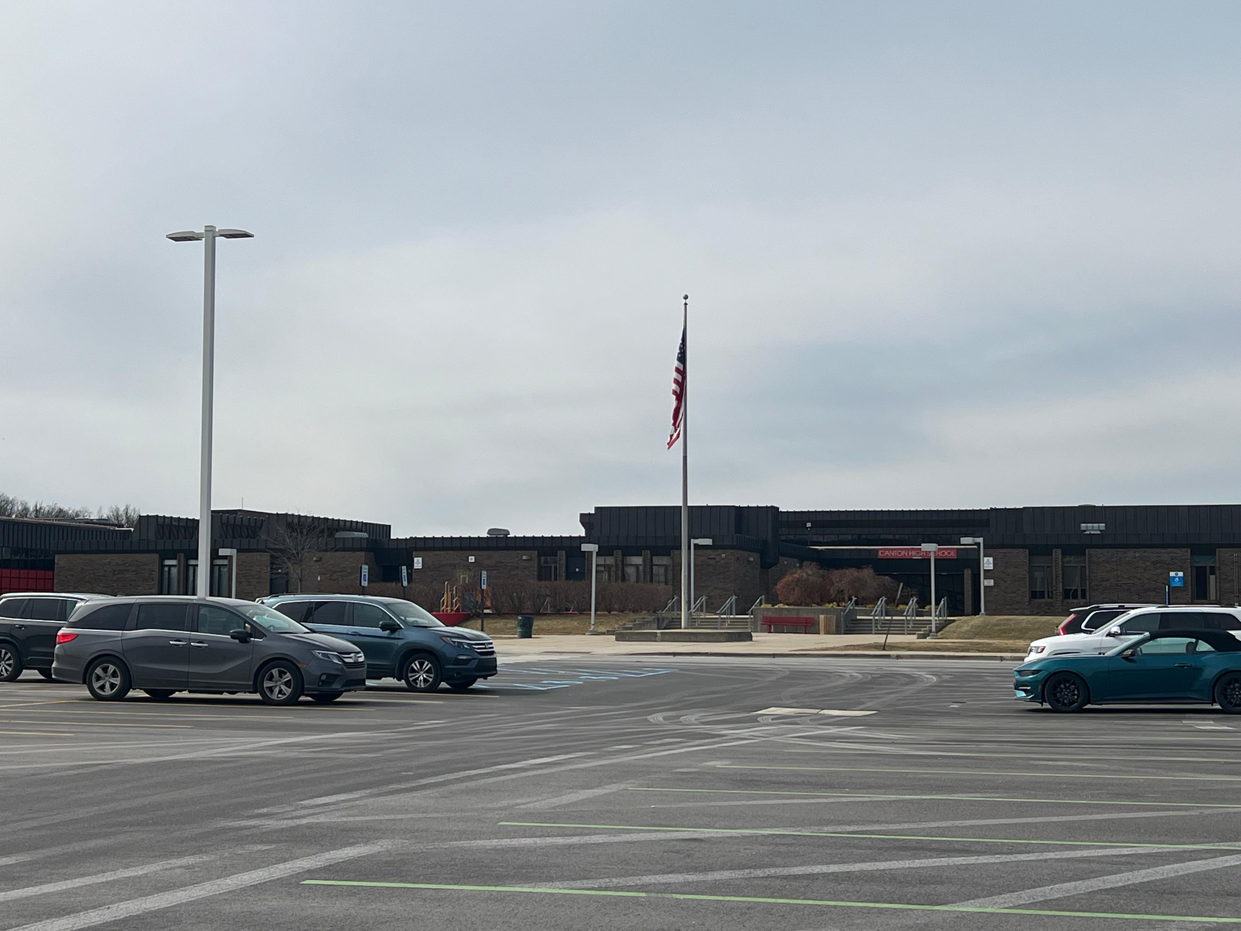 Low-rise brick school building with a flagpole and parking lot under an overcast sky.