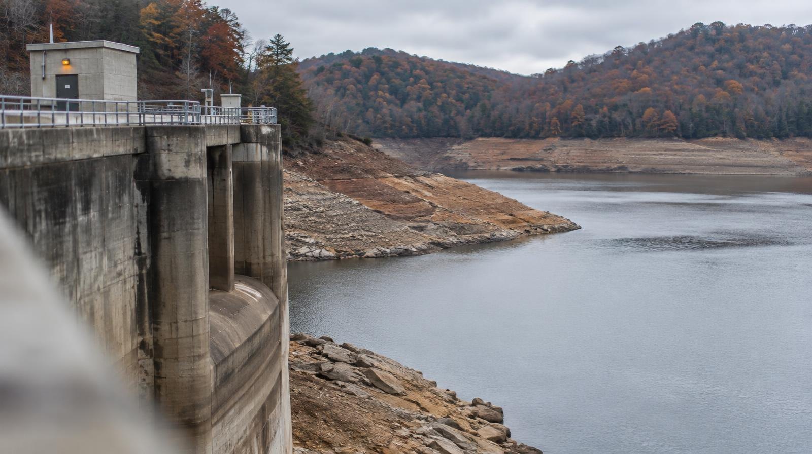 A concrete dam holding back a low reservoir with exposed rocky shoreline on an overcast day.