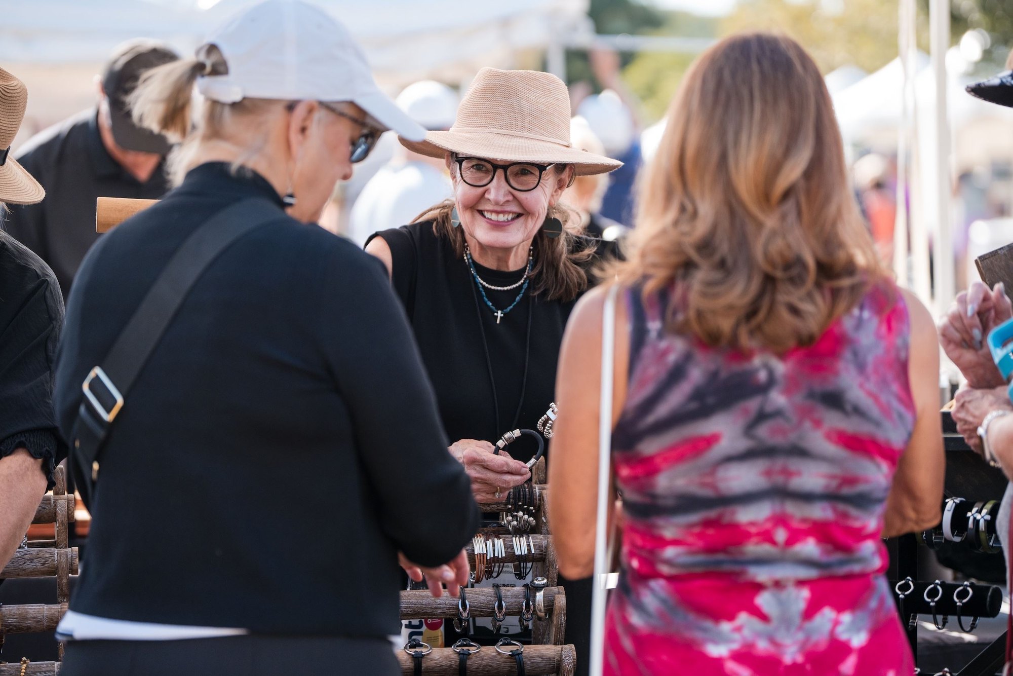 Three women browse items at an outdoor vendor booth under white tent canopies.
