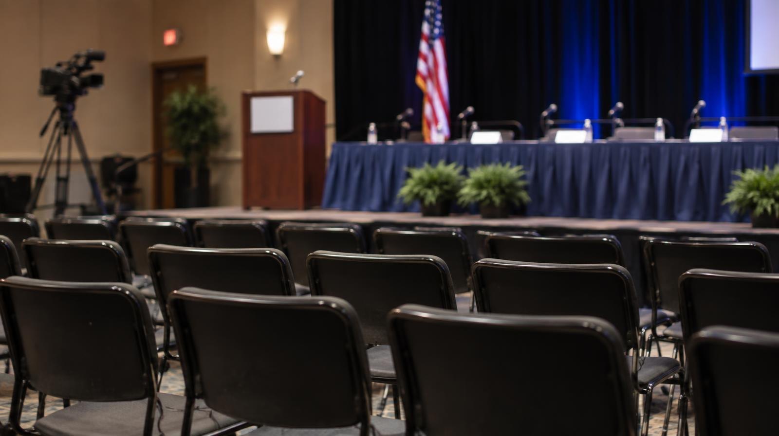 Rows of empty chairs face a raised dais in a public meeting room.