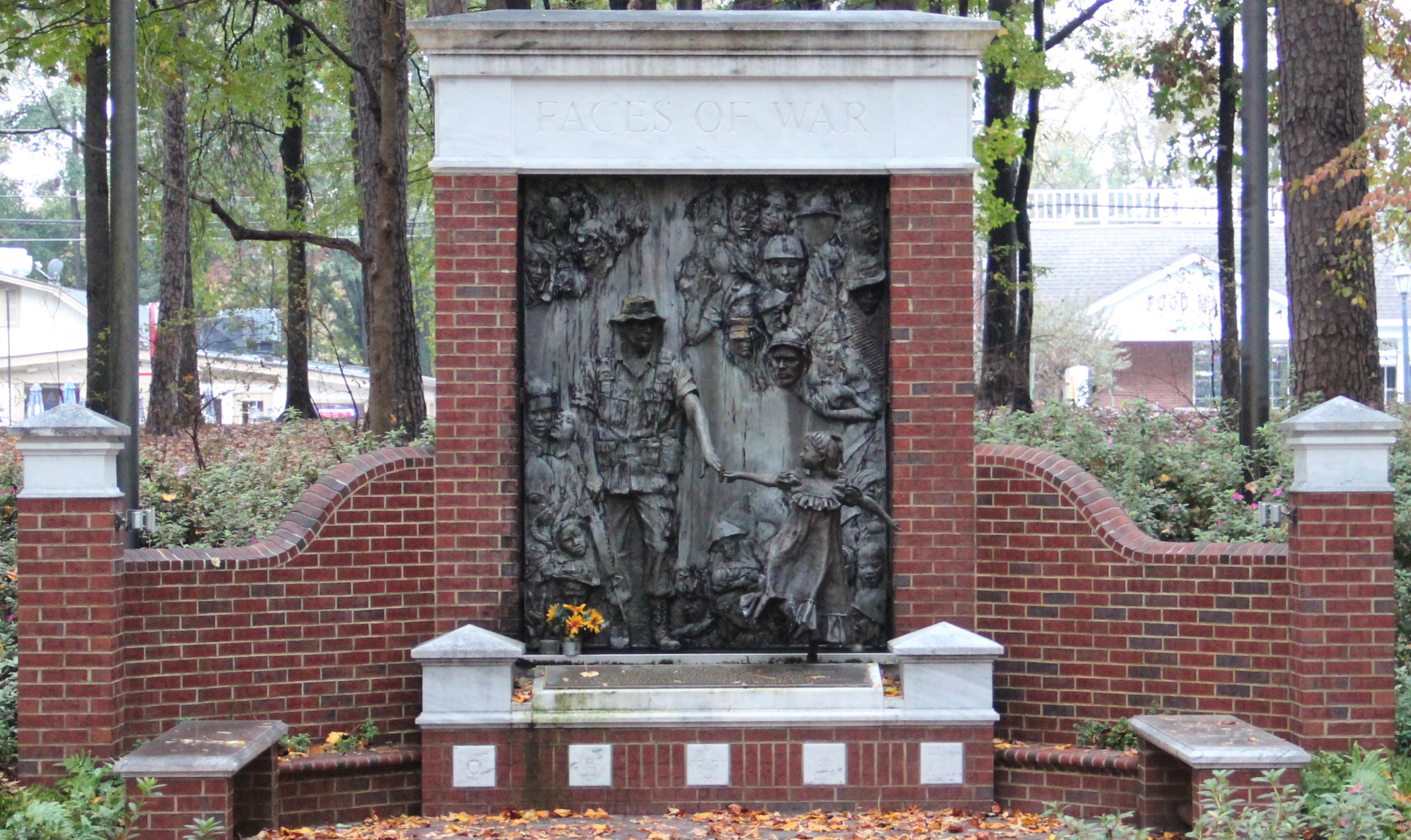 Bronze relief sculpture on a brick memorial flanked by curved walls and autumn trees.