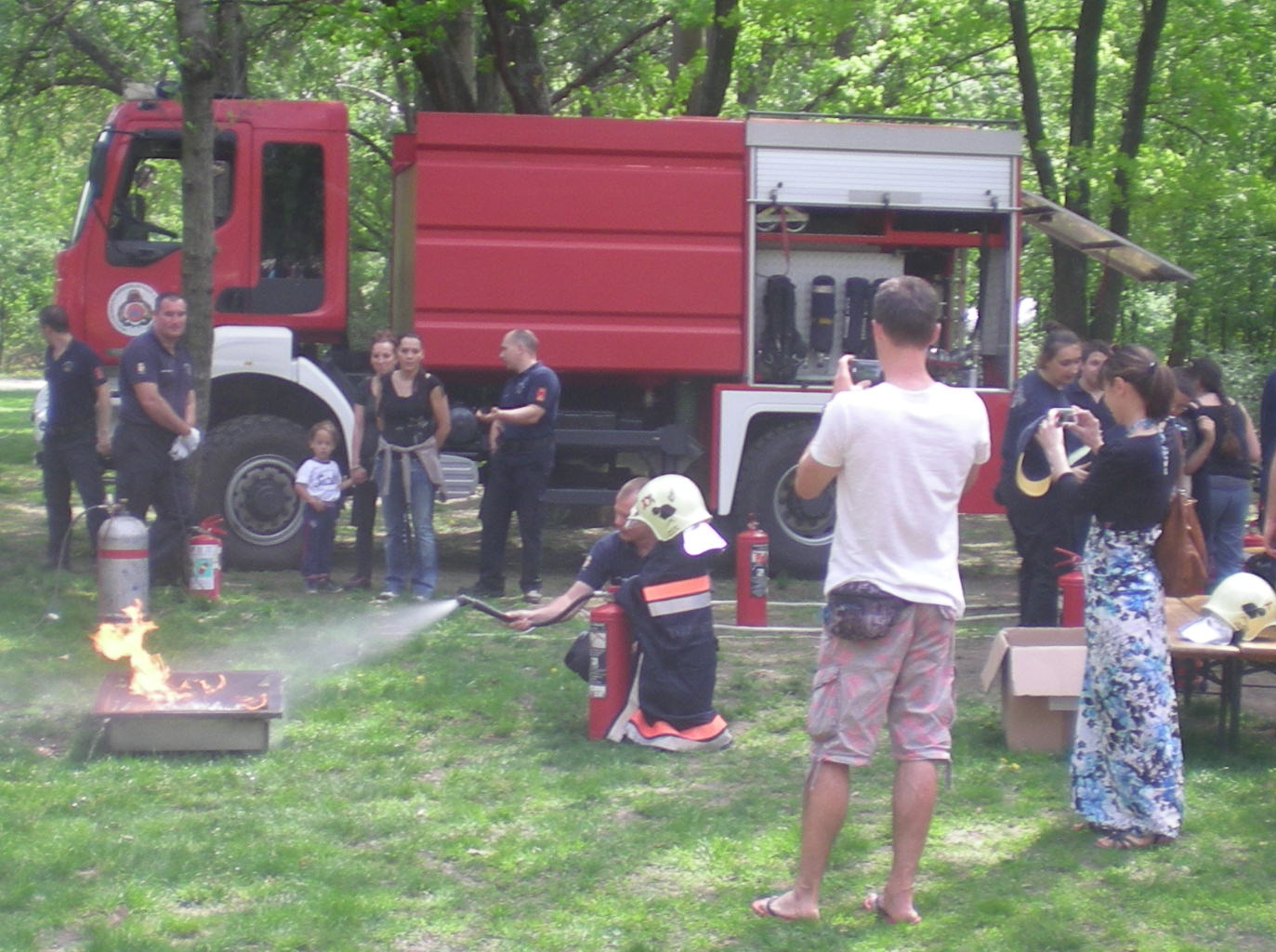 A child in a firefighter helmet and cloak aims a fire extinguisher, guided by a firefighter.