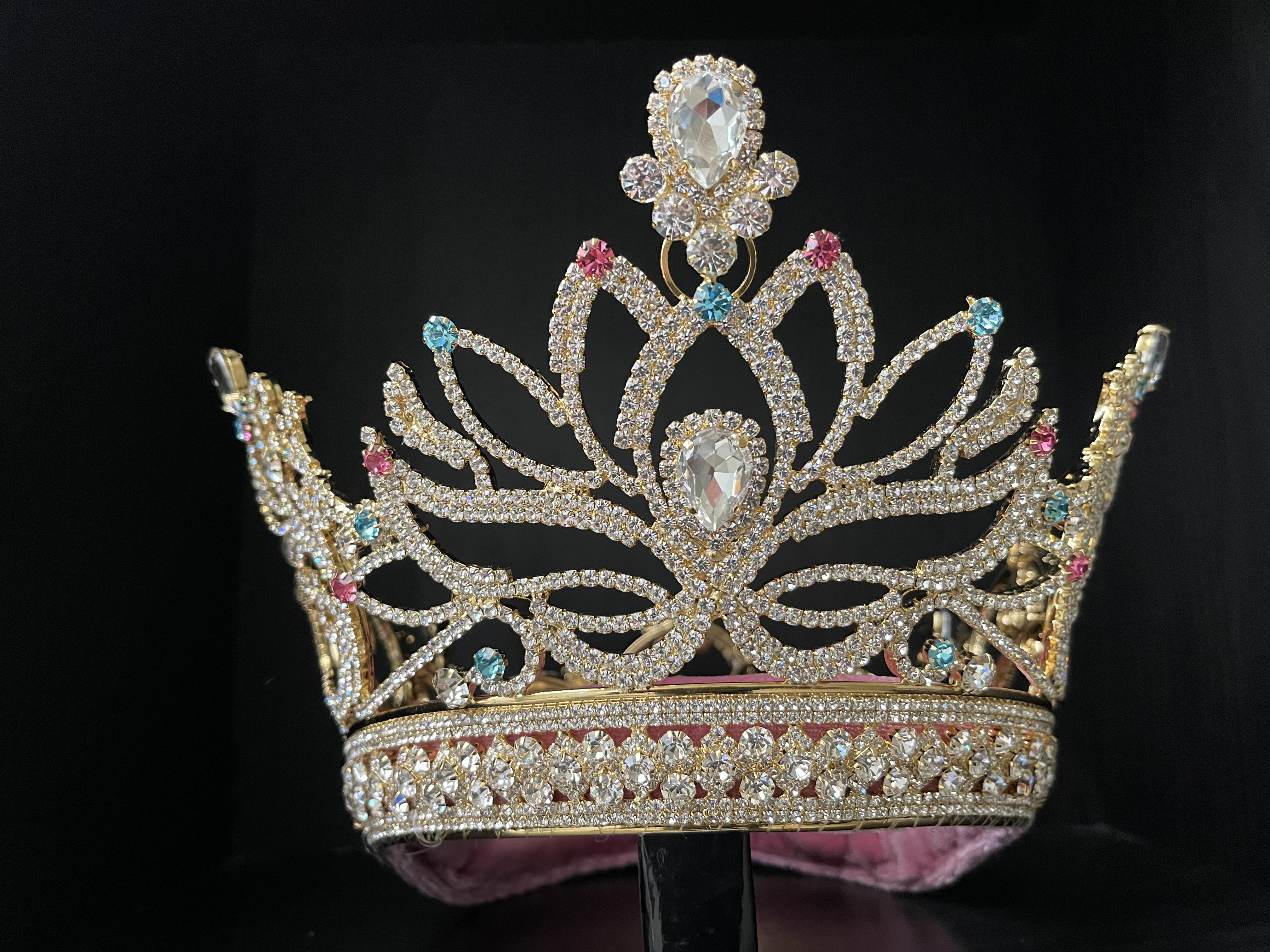 Ornate gold pageant crown with rhinestones and crystals on a dark background.