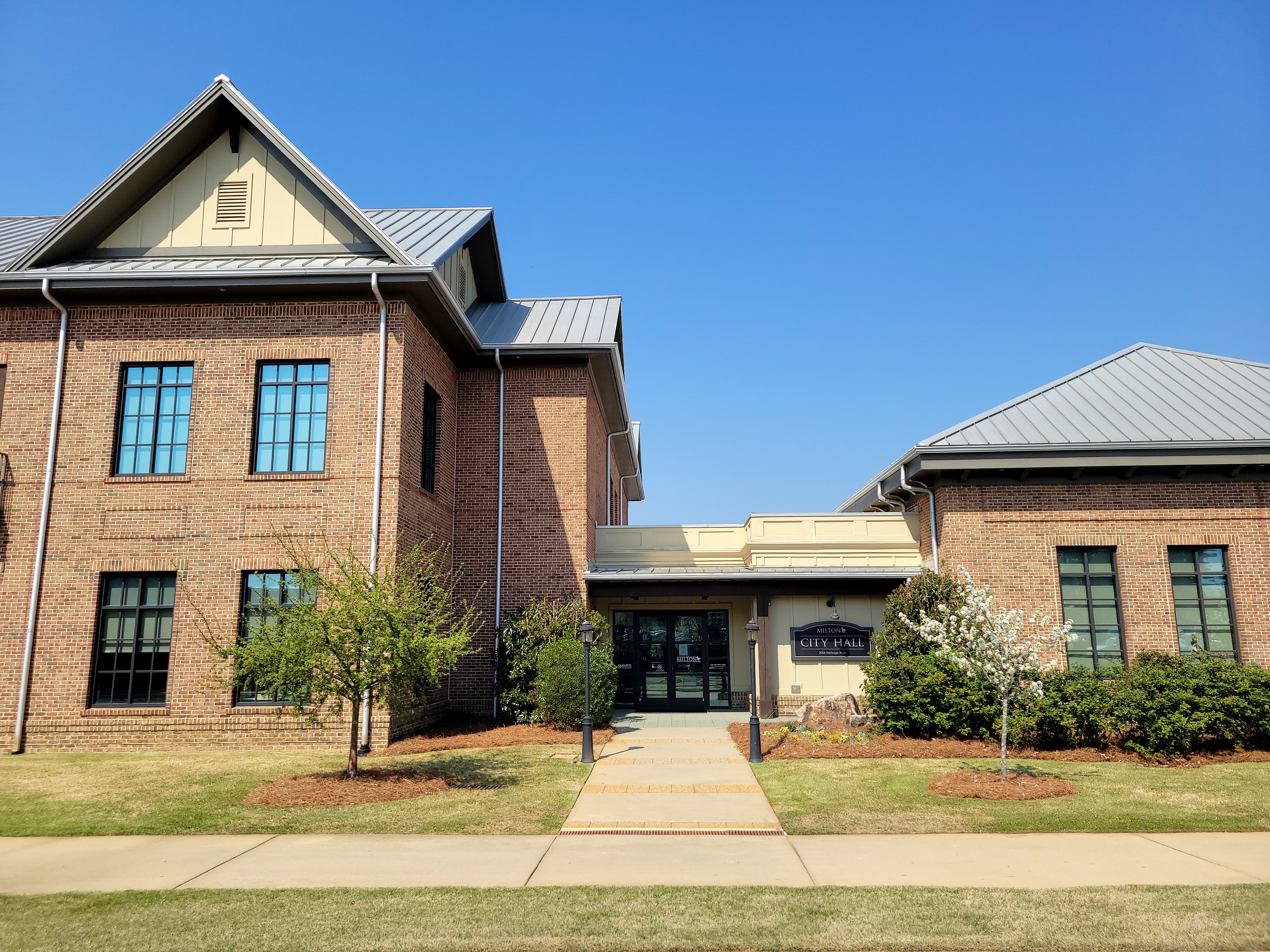 Two-story red brick city hall building with metal roof and landscaped entrance walkway.