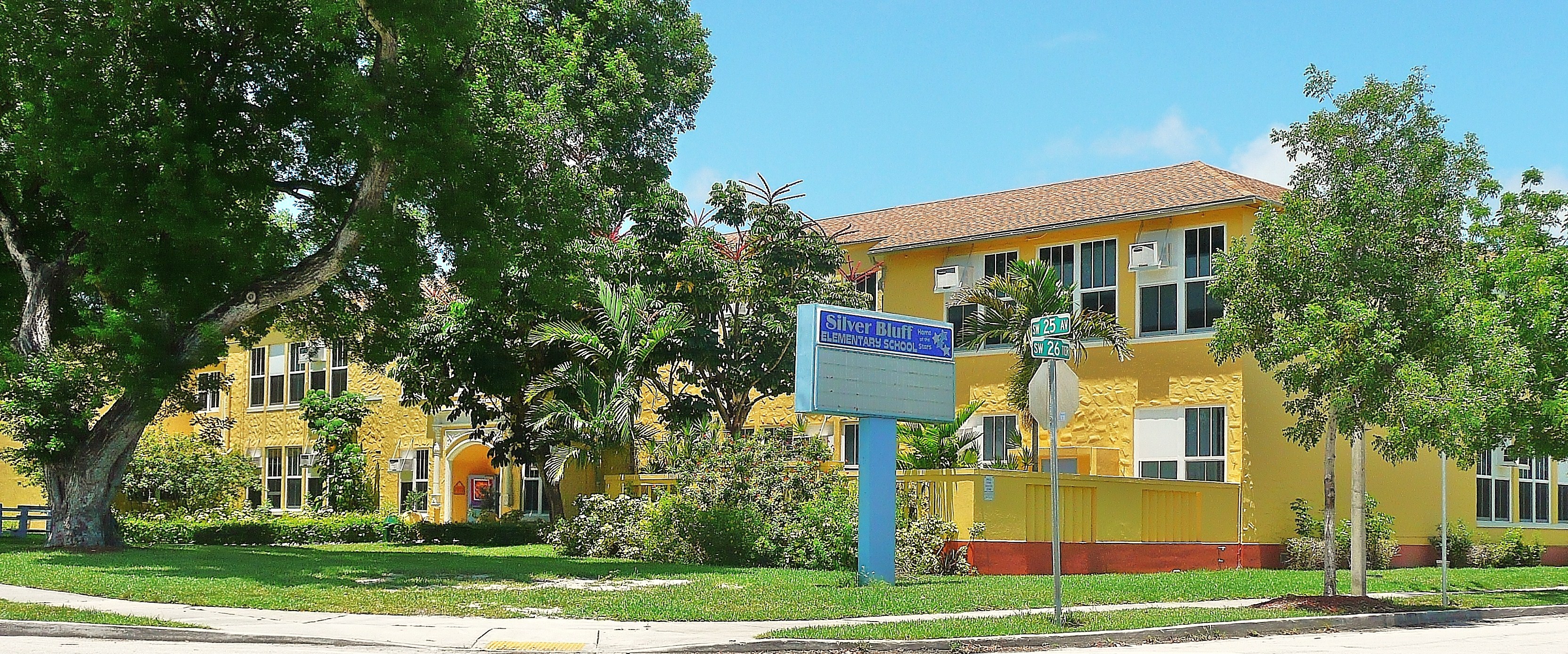 Two-story yellow school building with red tile roof and blue sign on a green lawn.