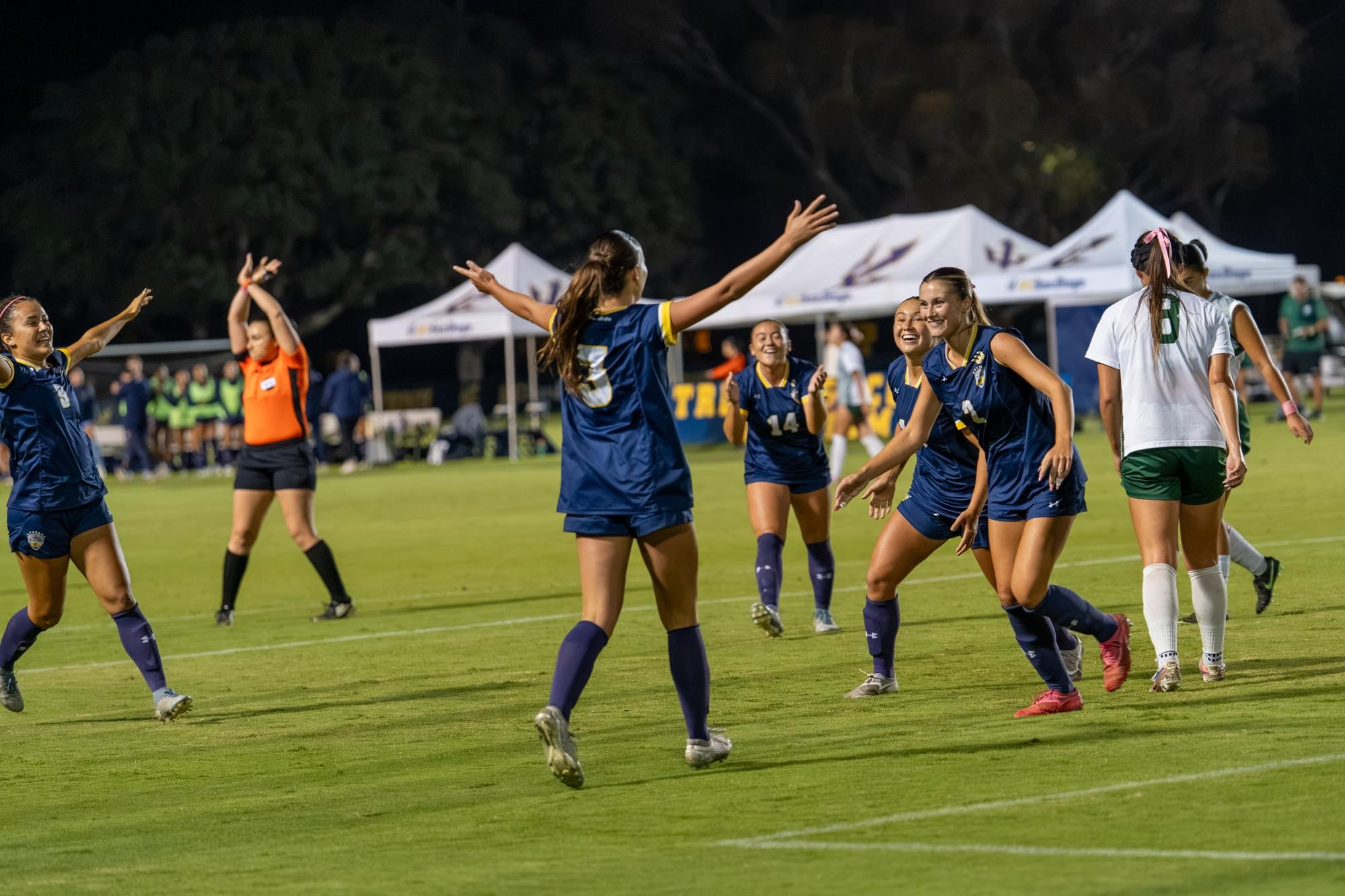 Women's soccer players in navy uniforms celebrate on a lit field at night.