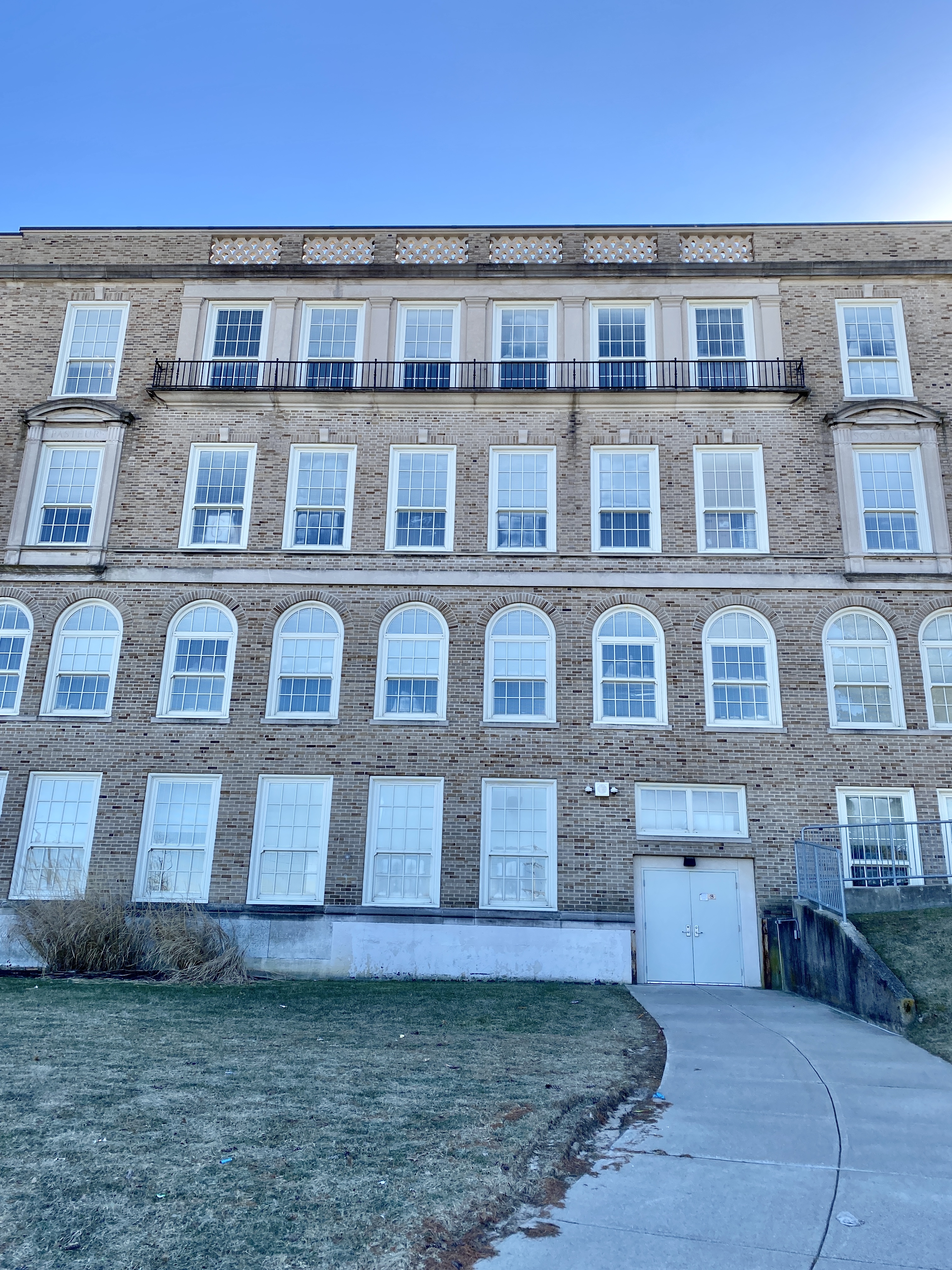 Multi-story buff brick school building with arched windows and a concrete walkway leading to the entrance.