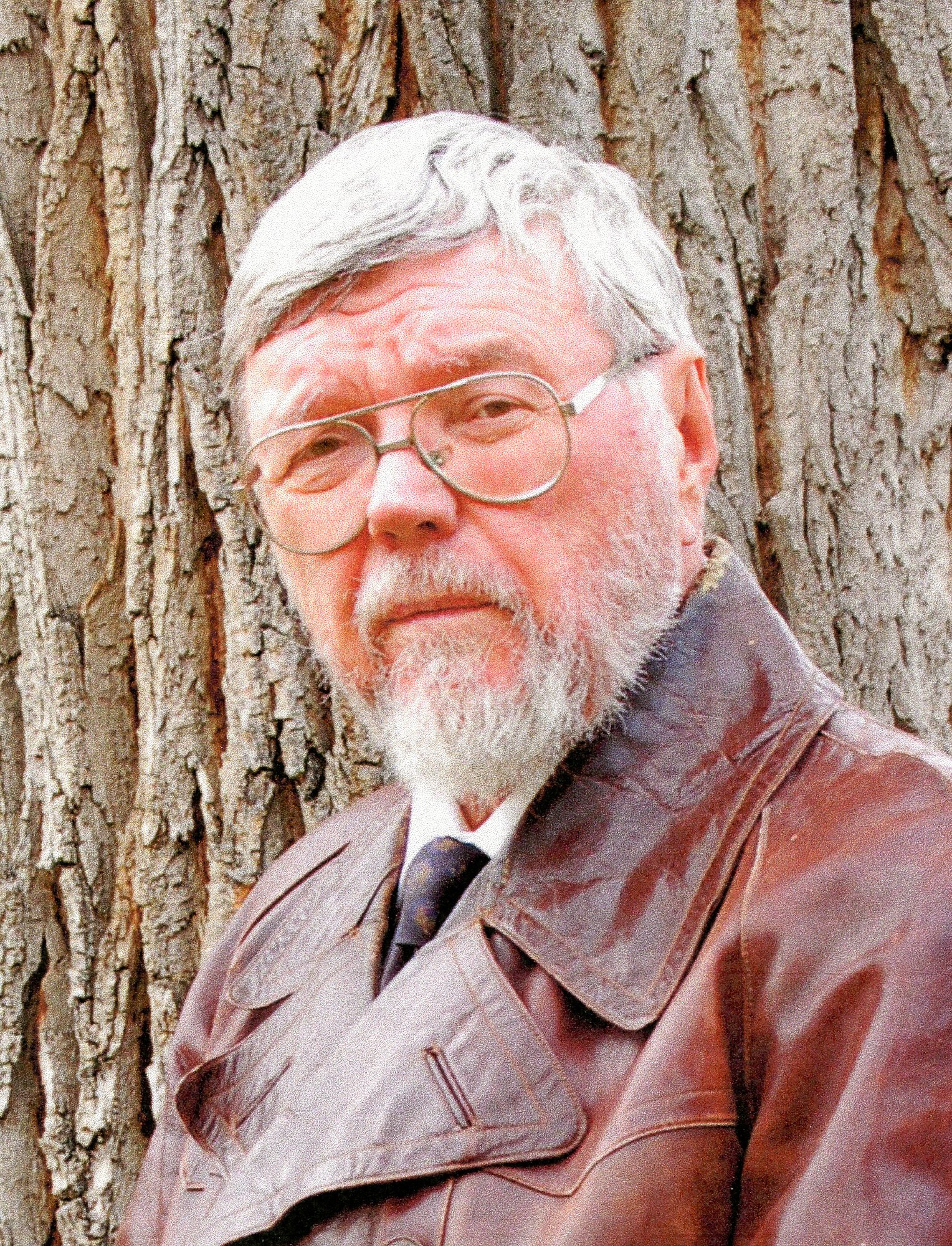 Older white-haired man with a gray beard and glasses in a brown leather jacket, standing outdoors before a large tree.