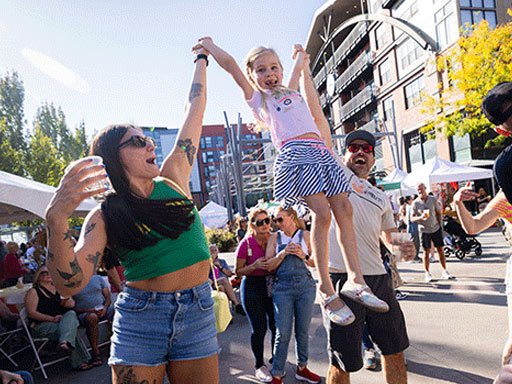 Two adults lift a young girl by her hands at a sunny outdoor street festival with vendor tents.