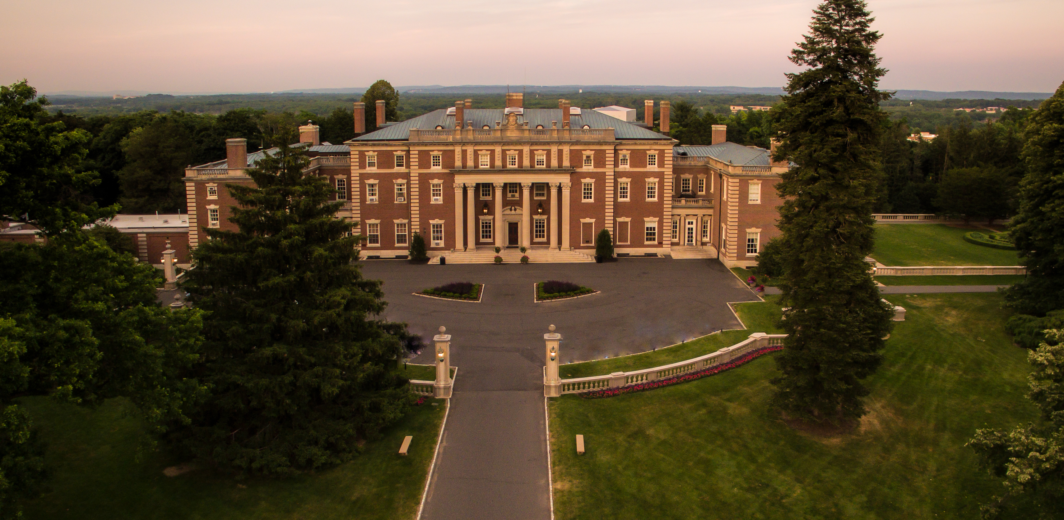 Aerial view of a large brick mansion with columned portico and circular driveway at dusk.