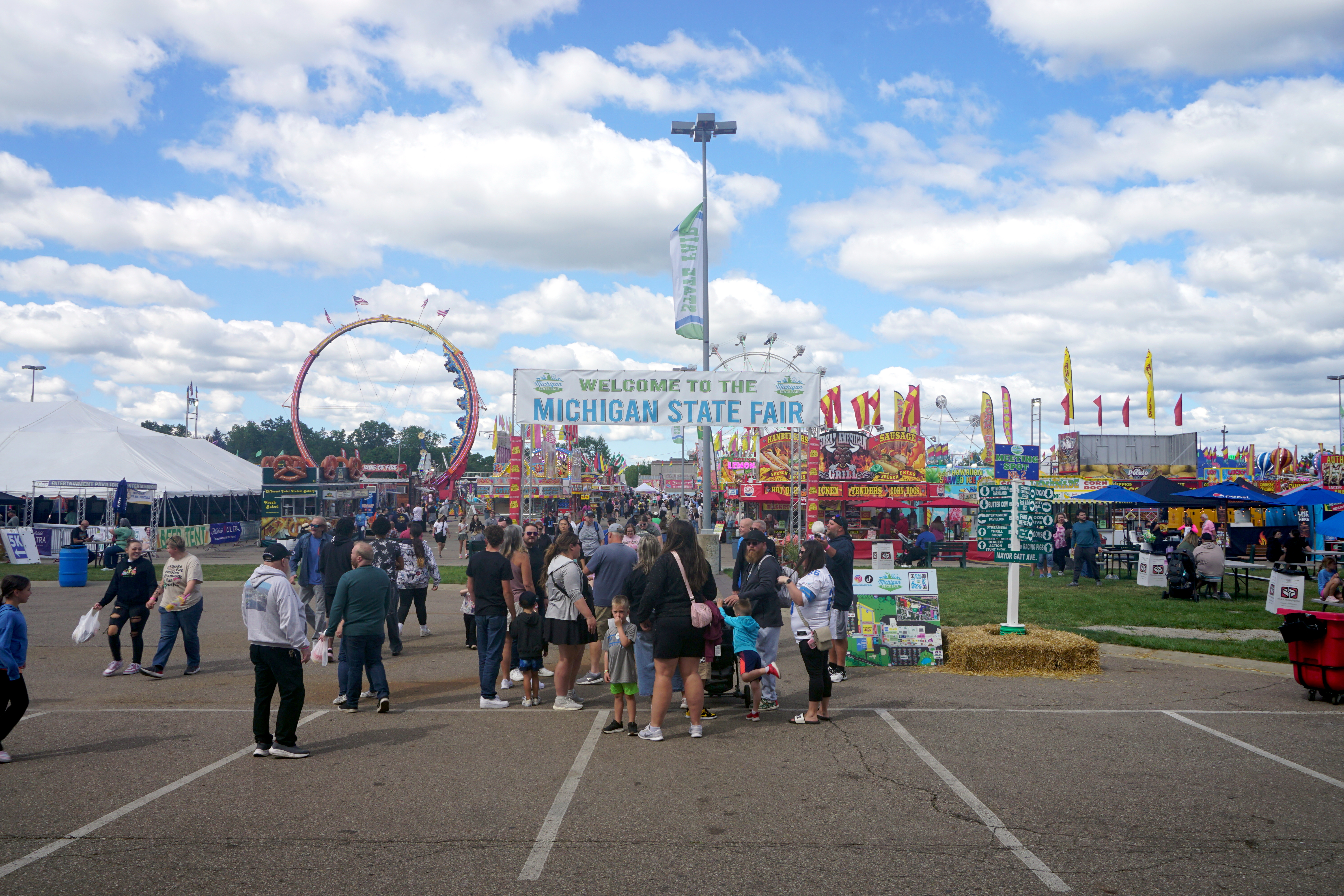 Outdoor fair midway with a Ferris wheel, carnival rides, colorful tents, and crowds of visitors.