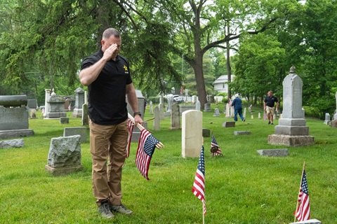 A man in a black polo shirt salutes at a gravestone while holding small American flags.