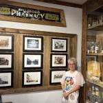 A woman in a white t-shirt stands in front of framed historical photos and a vintage pharmacy sign.