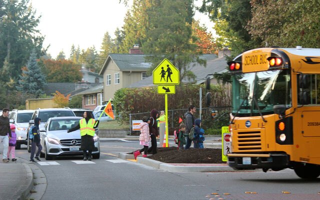 Crossing guard in yellow vest stops a car as children cross in front of a school bus.