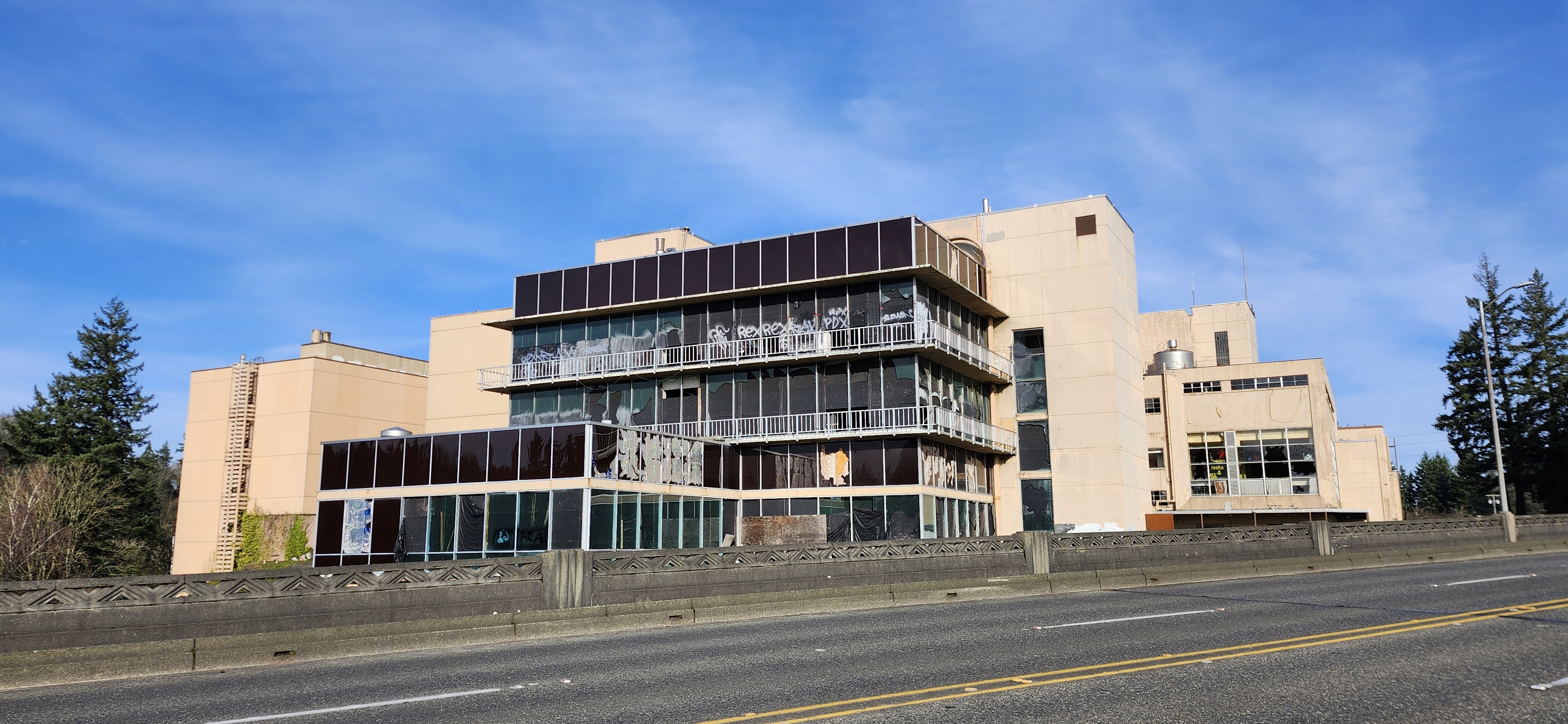 Large multi-story industrial building with broken windows and graffiti, set beside a road.