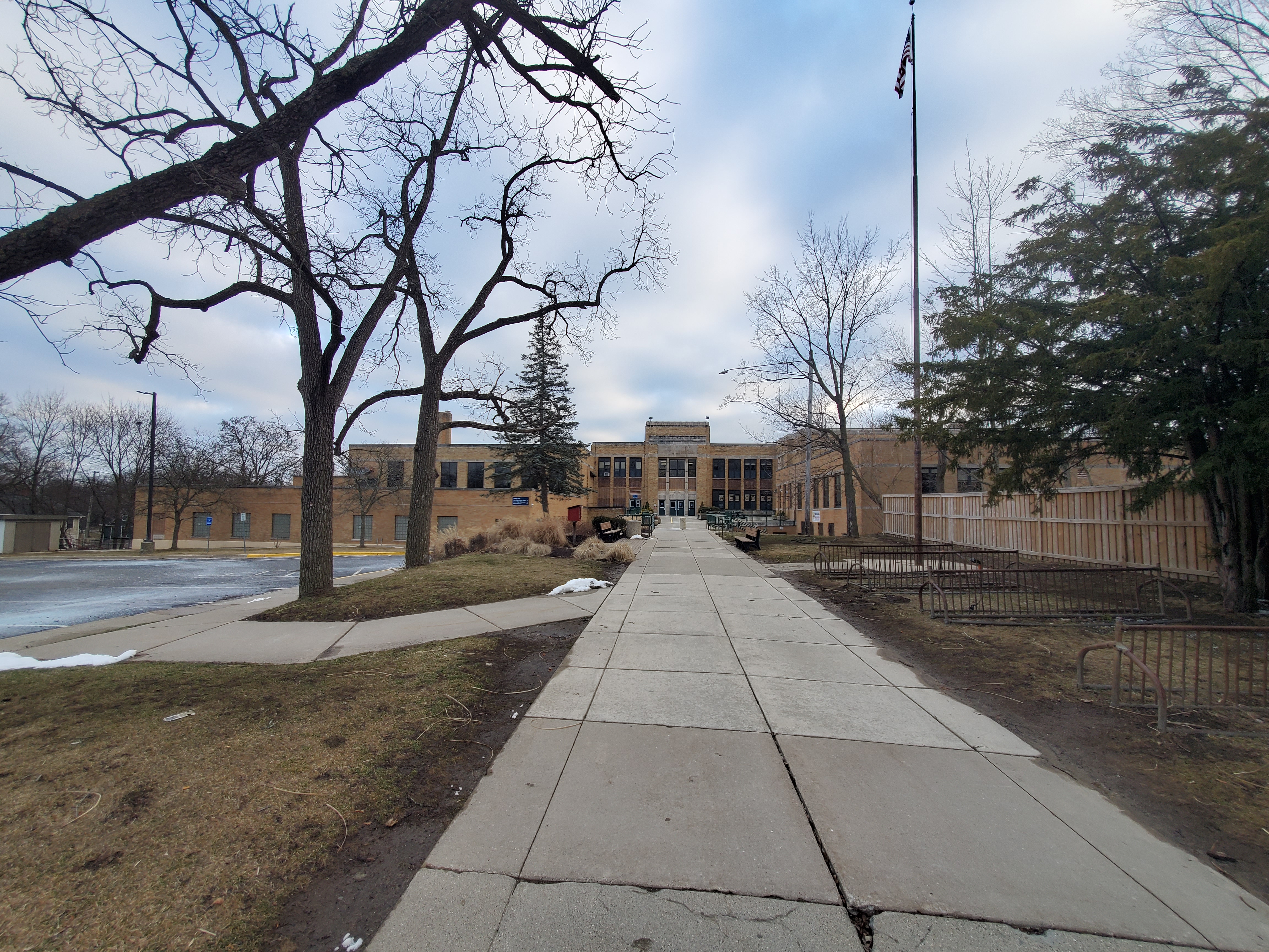 Brick school building entrance with a wide walkway, flagpole, and bare trees in winter.