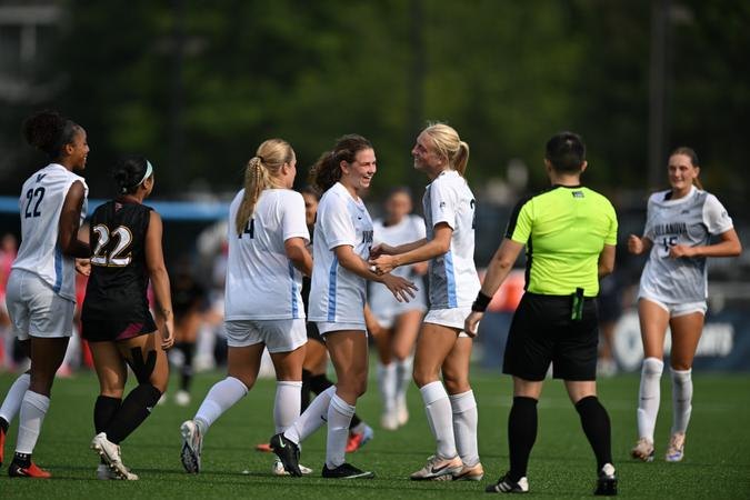 Women's soccer players in white uniforms celebrate on a green field.
