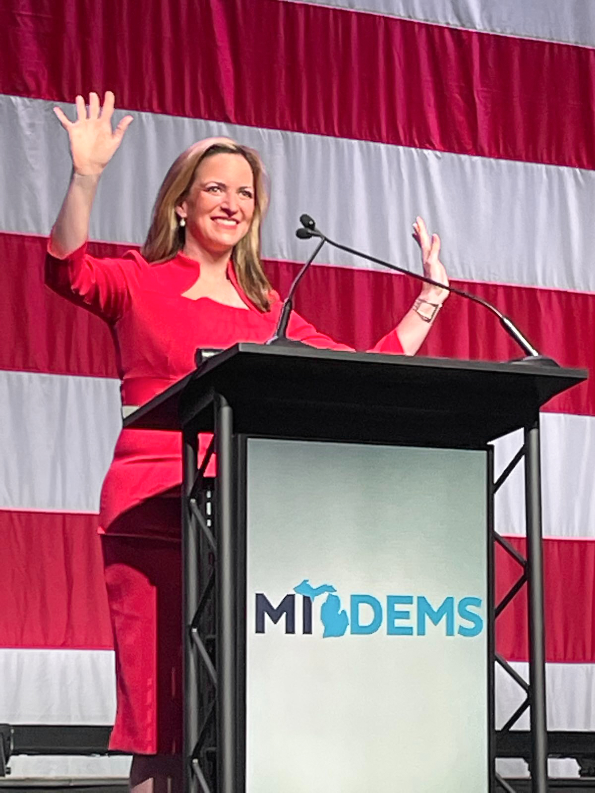 Woman in red outfit waving and smiling at a podium with an American flag backdrop.