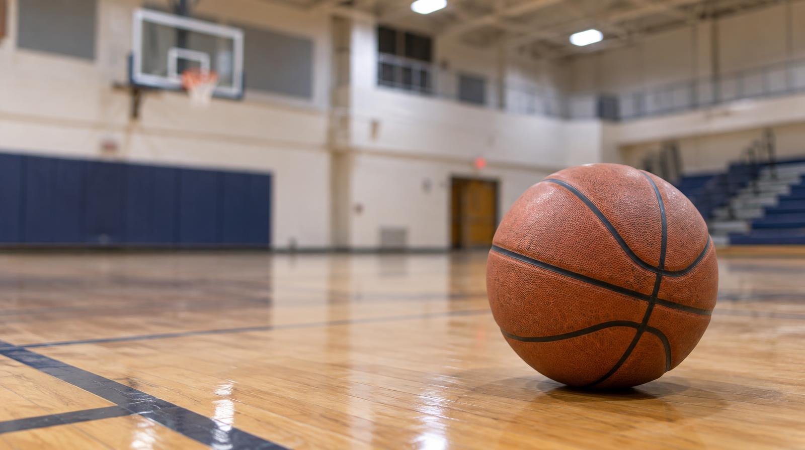 Empty indoor gymnasium with hardwood basketball court and hoop under bright overhead lights.