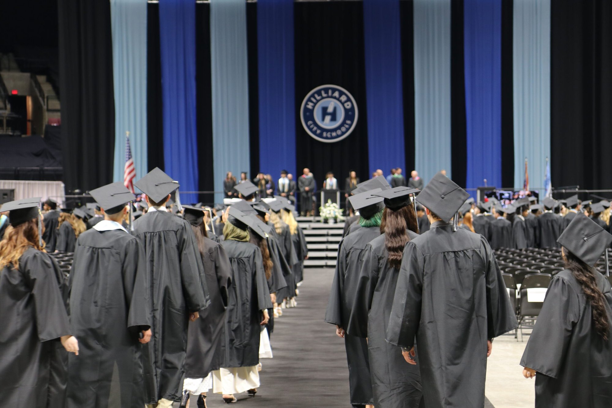 Students in black graduation caps and gowns walk toward a stage in an arena.