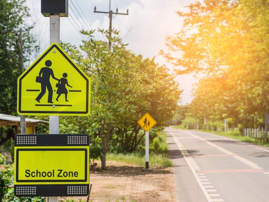 Yellow school zone crossing sign with pedestrian figures along a tree-lined road.