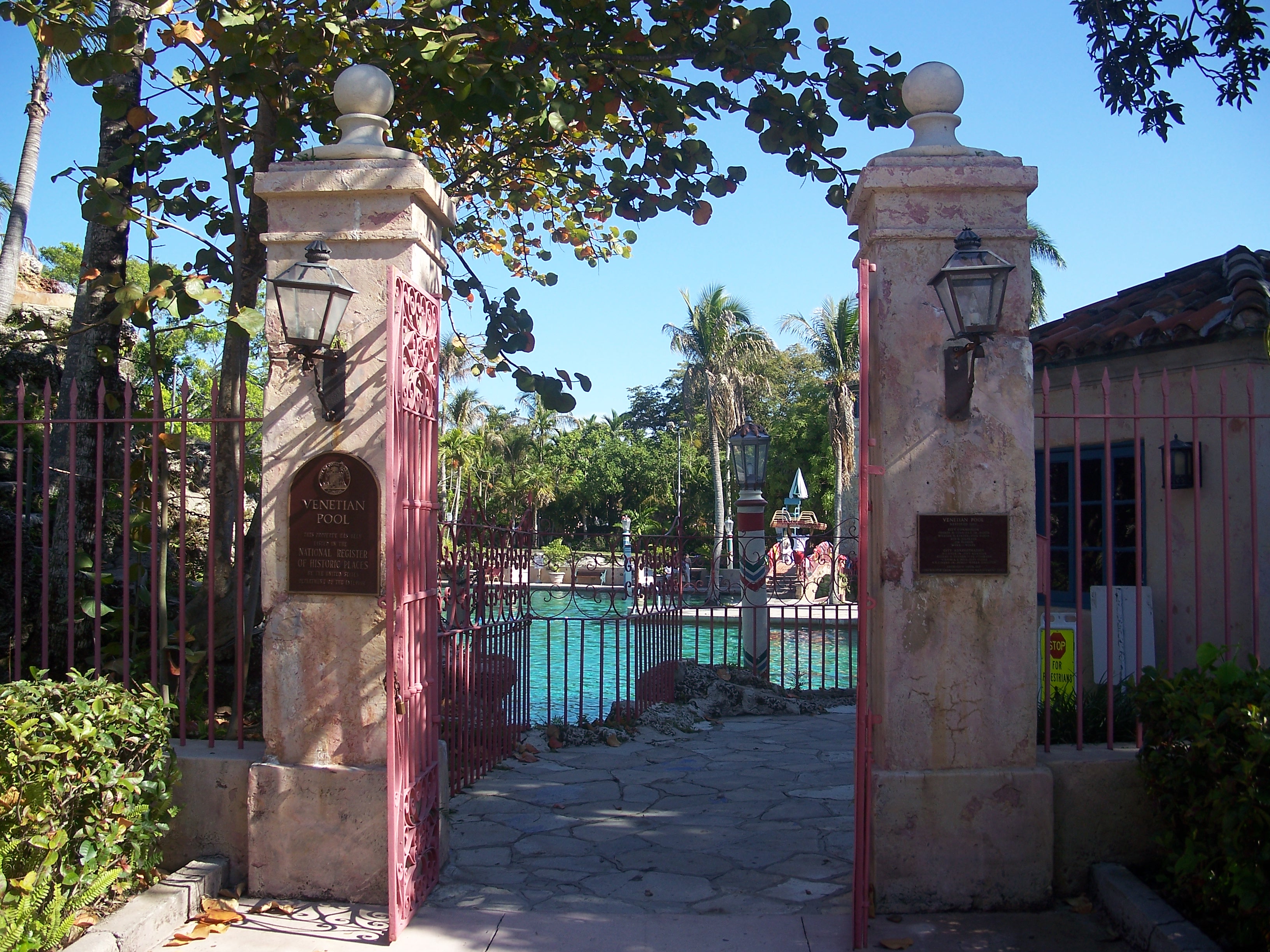Ornate red iron entrance gate between stone pillars, with a turquoise pool and palm trees visible beyond.