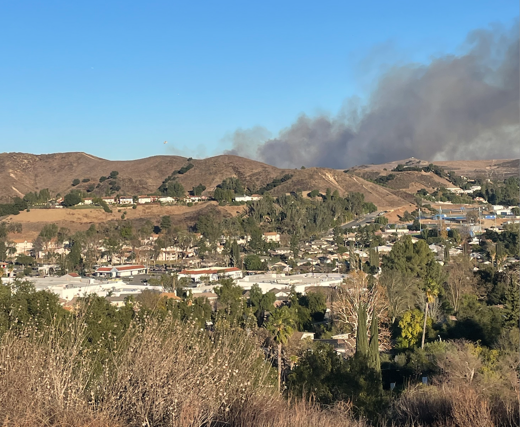 Dark smoke column rising behind a dry hillside above a residential neighborhood.