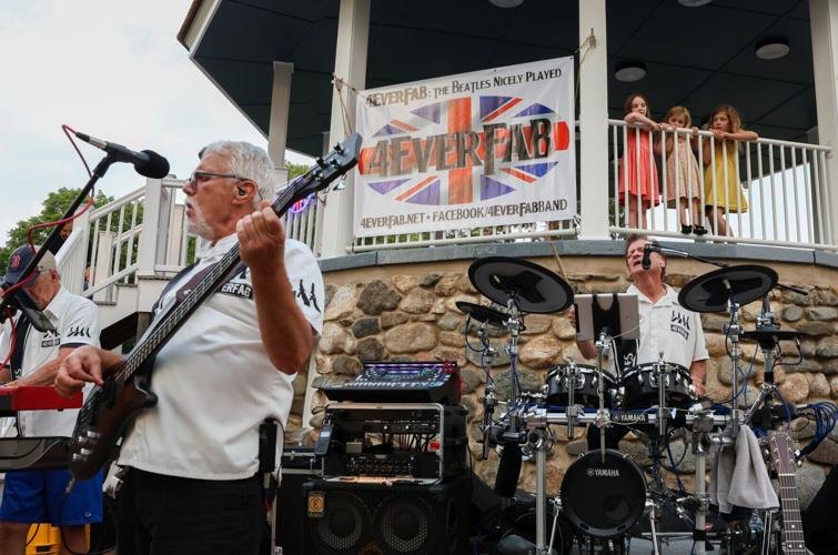 A guitarist and drummer perform outdoors under a gazebo banner at a summer concert.