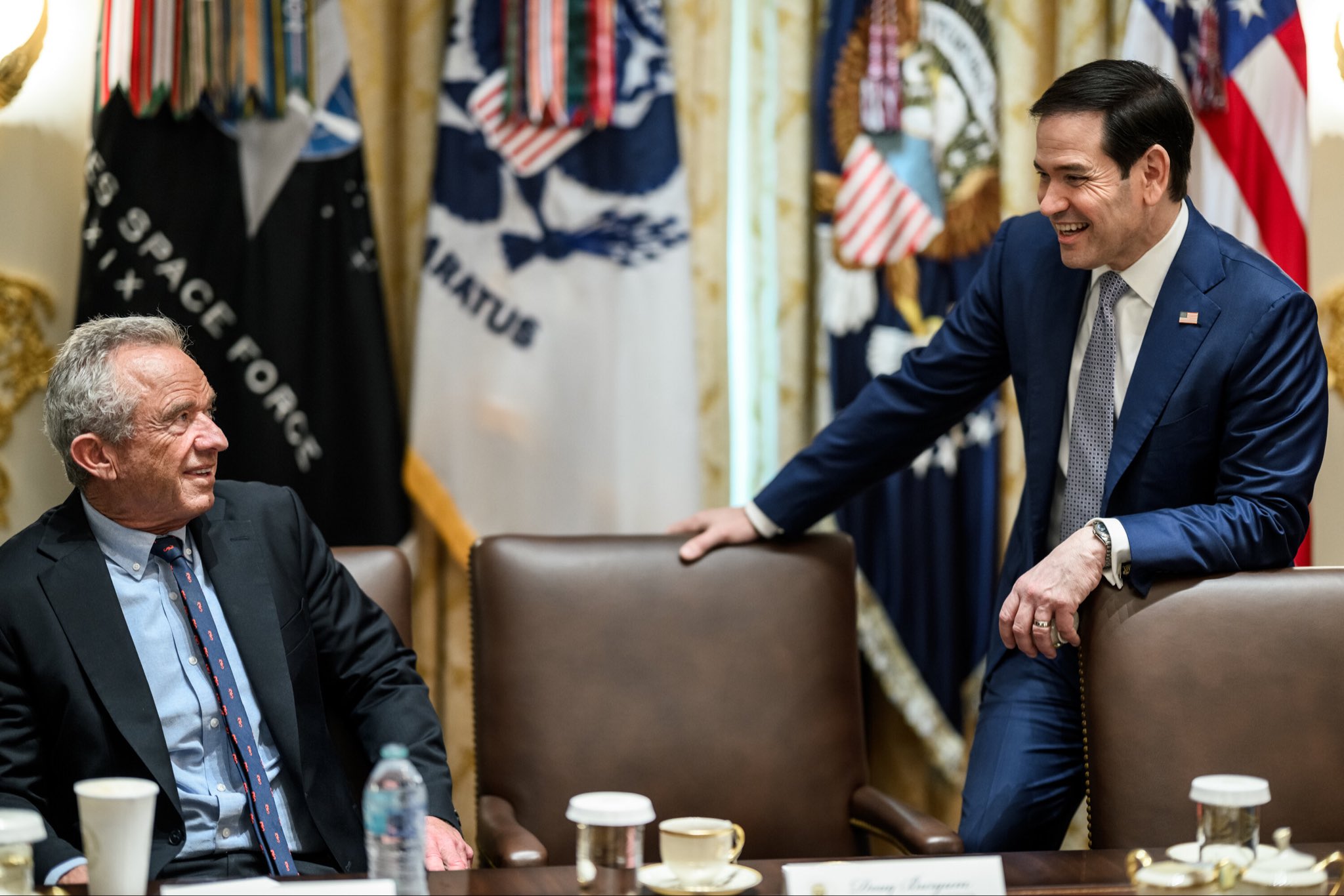 Two men in suits at a conference table with American flags in the background.