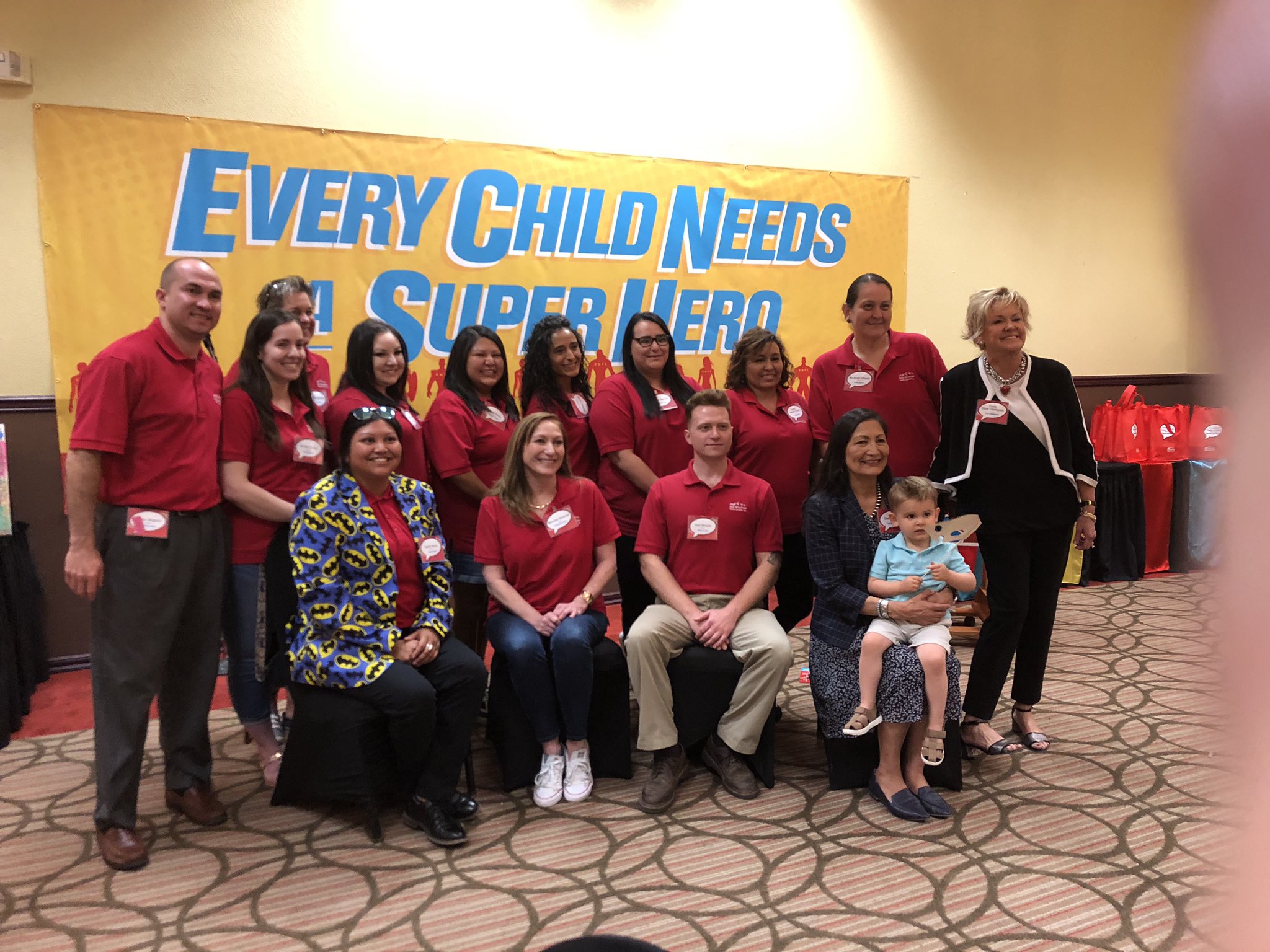 Group of people in red polo shirts pose in front of a yellow superhero banner at a foster care event.