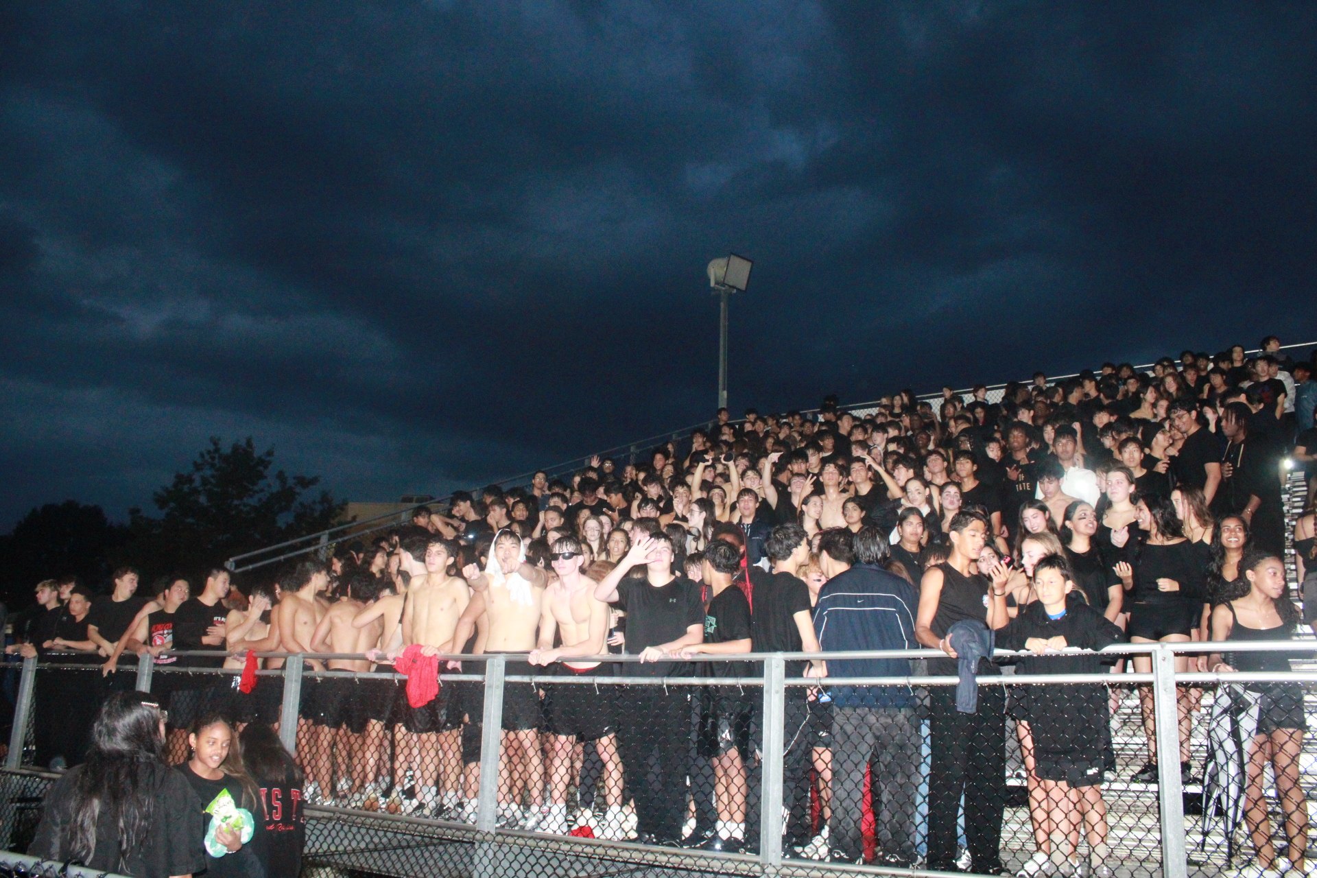 Dense crowd of students in black shirts filling bleachers under a dark, overcast night sky.