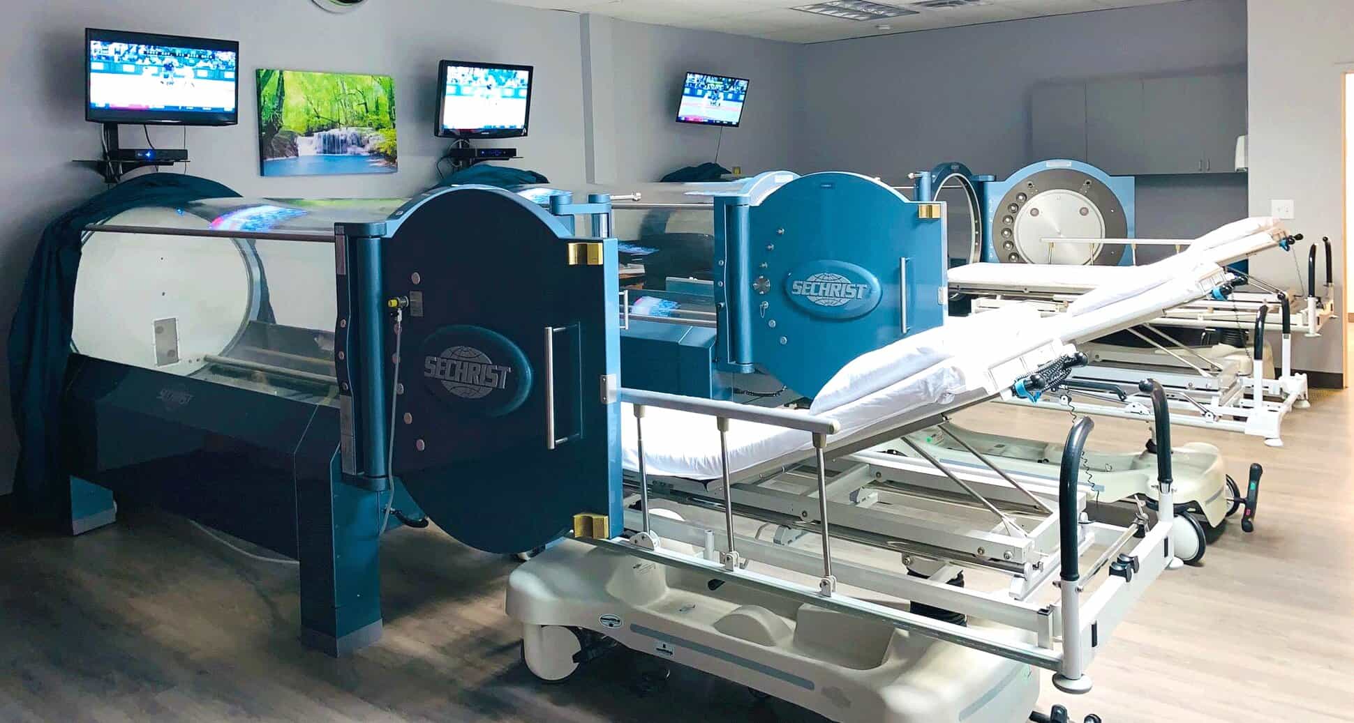 Three blue cylindrical hyperbaric oxygen chambers in a clinical room with hospital beds.