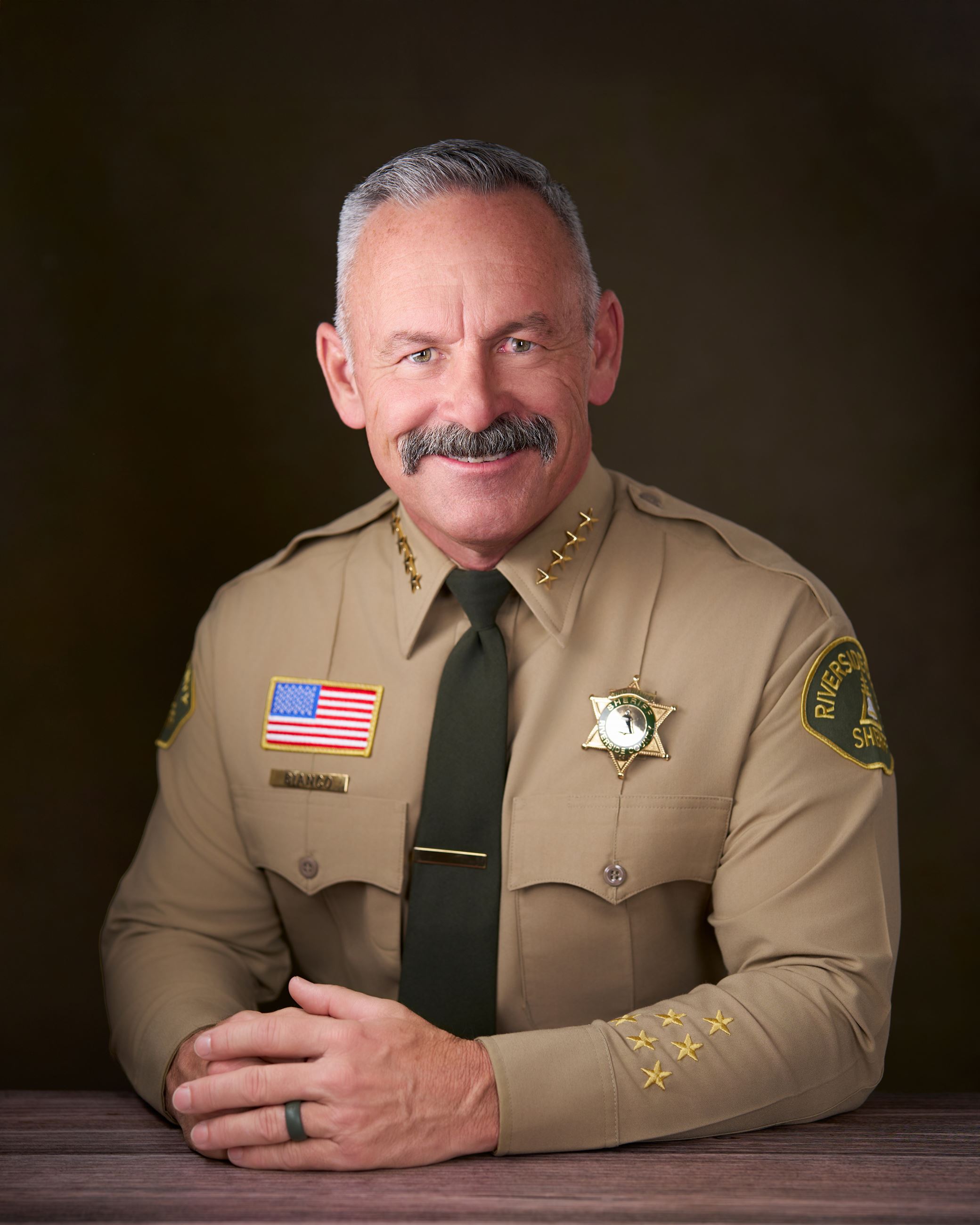 A man in a tan sheriff's uniform with a gold badge and four gold stars, seated with arms crossed.