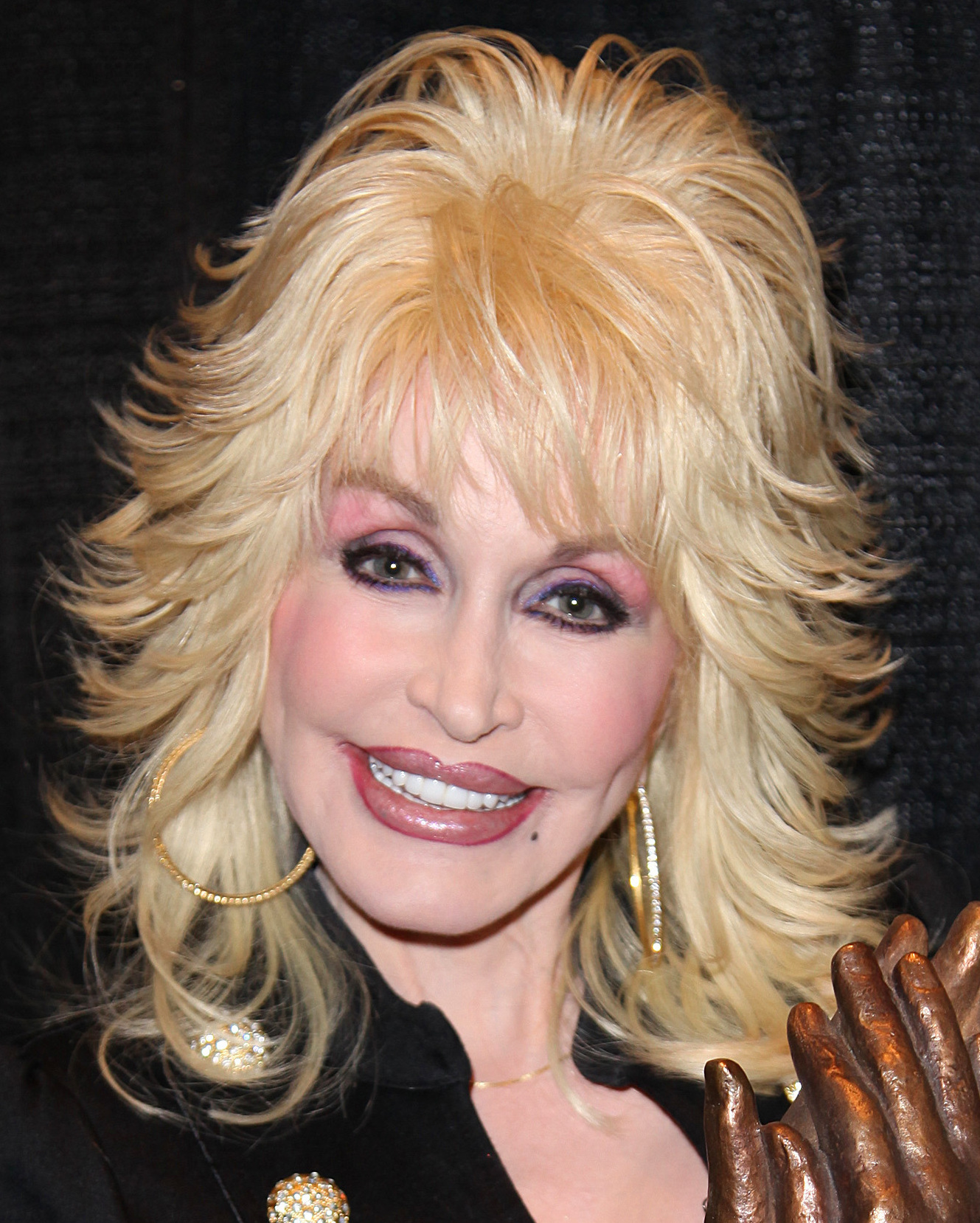 Woman with voluminous blonde hair and gold earrings smiles at an indoor event.