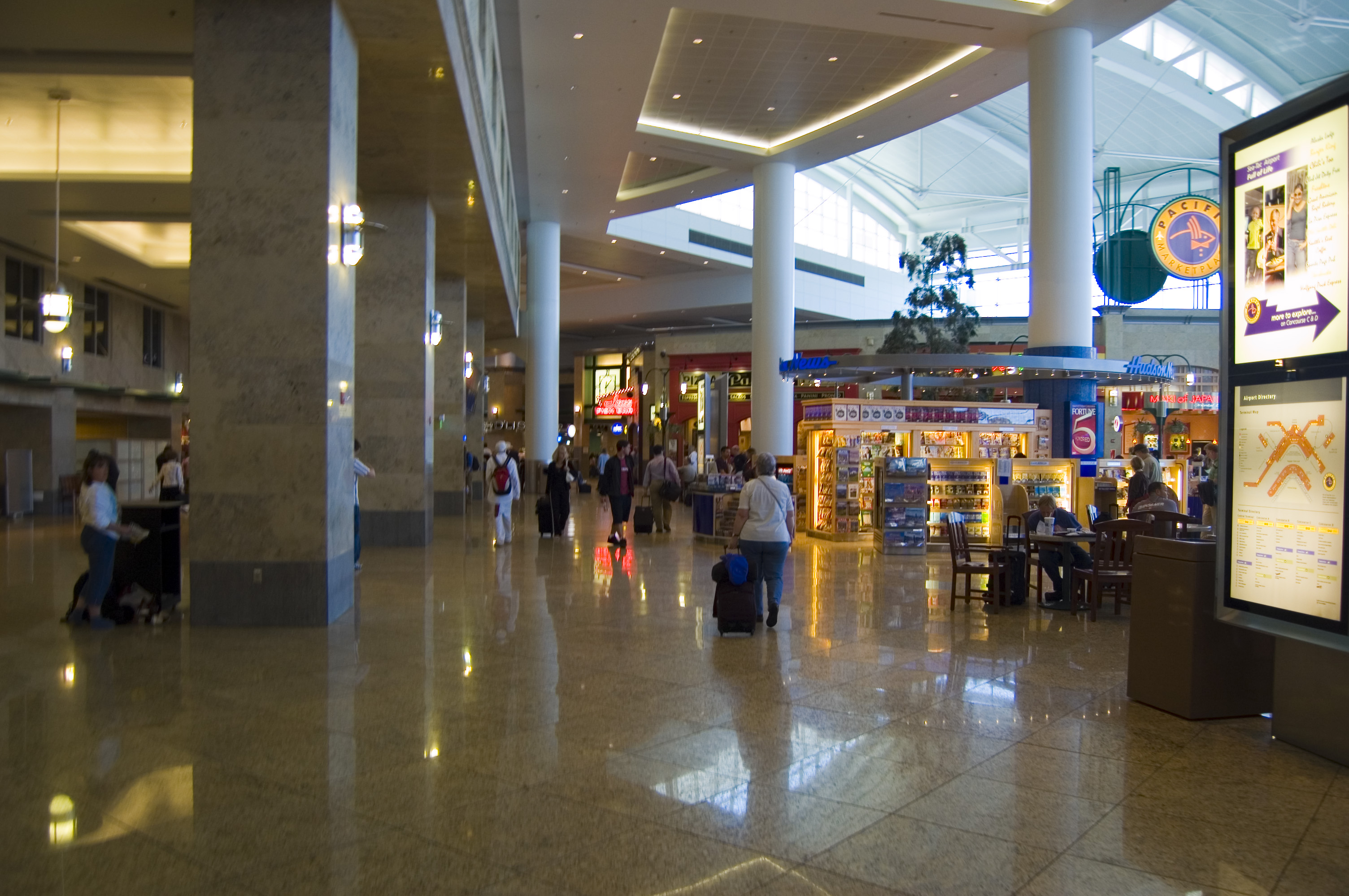 Travelers walk through a wide airport terminal lined with food and retail shops.