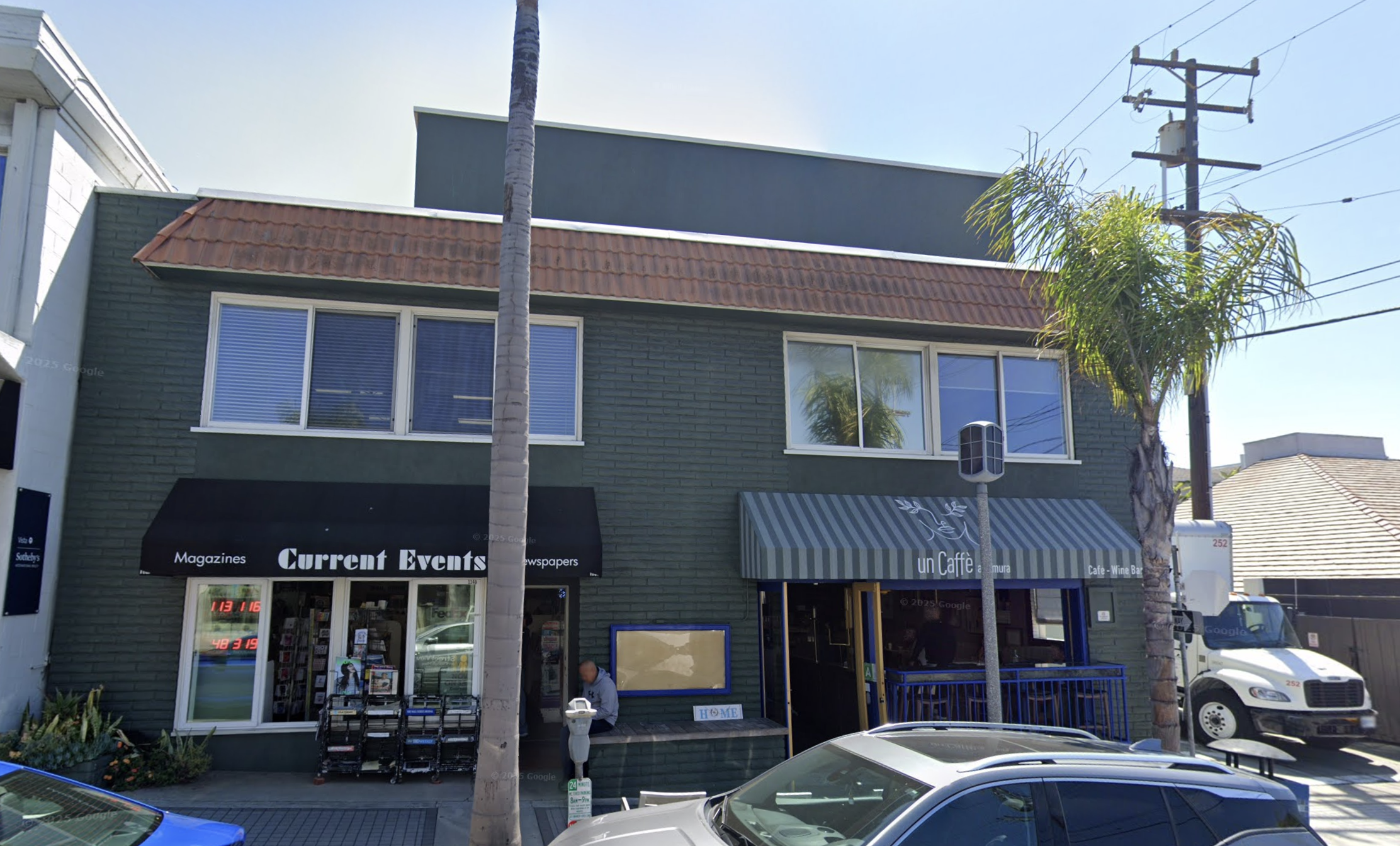 Two-story green brick building with Current Events newsstand and a café at street level, framed by palm trees and parked cars.