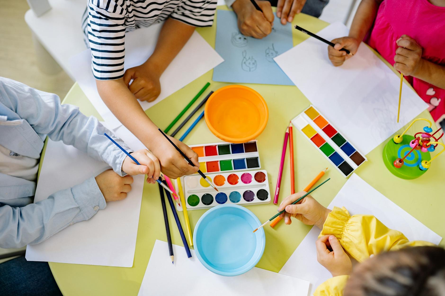 Children's hands reaching over a table with watercolor paints, colored pencils, and paper drawings.