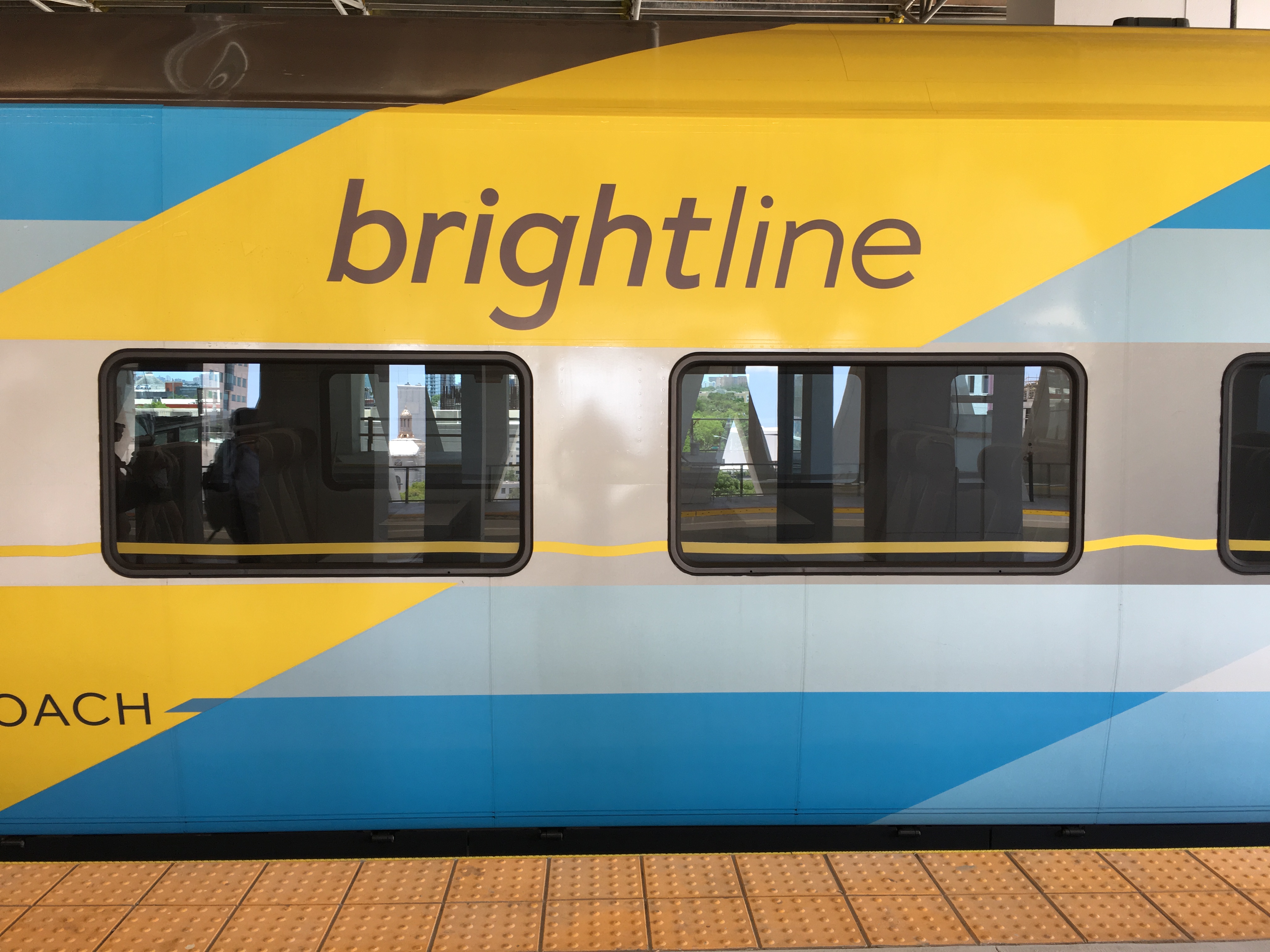 Side of a Brightline passenger train car with yellow, blue, and white livery at a station platform.