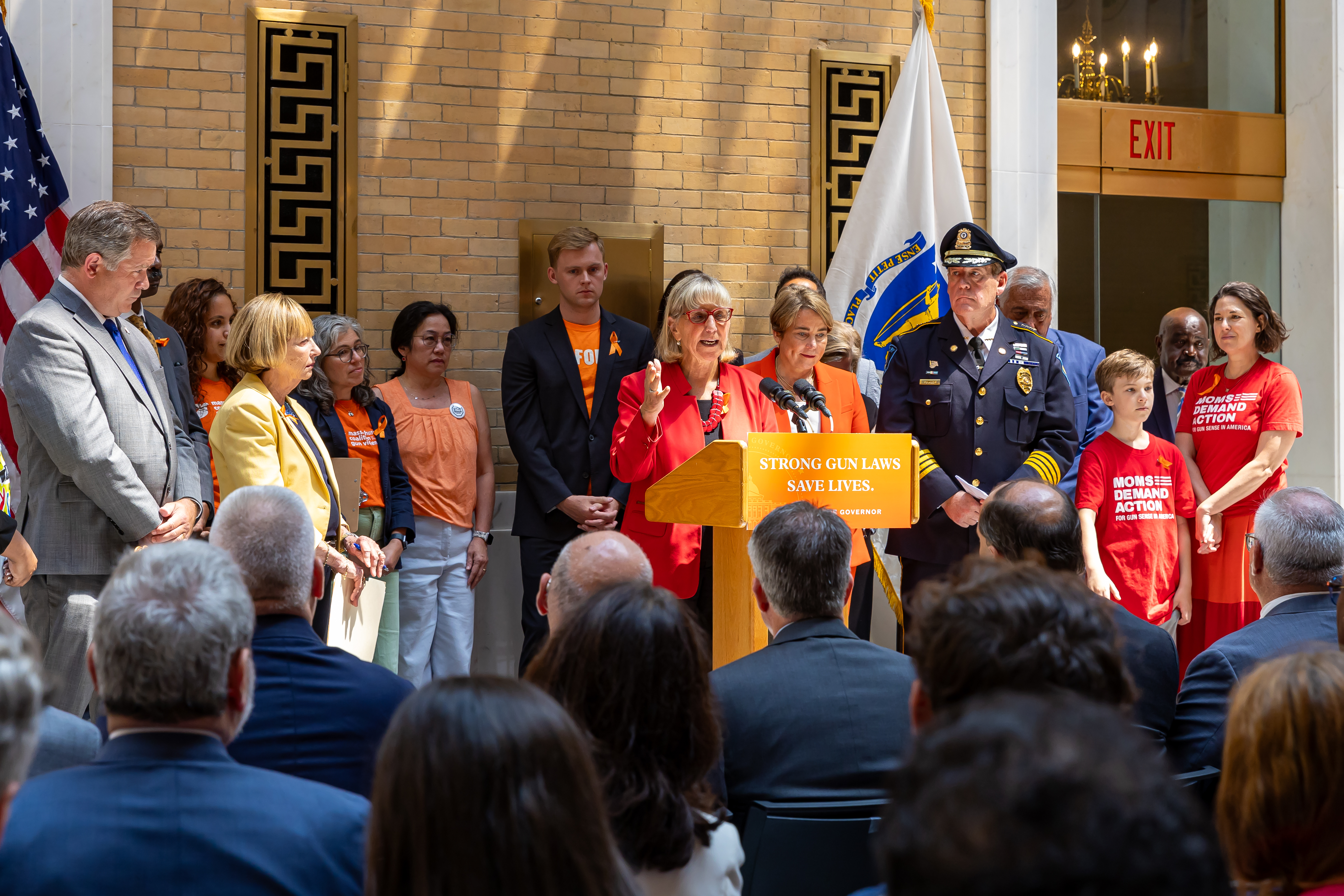 A crowd of officials and advocates stands at a podium inside a government chamber.