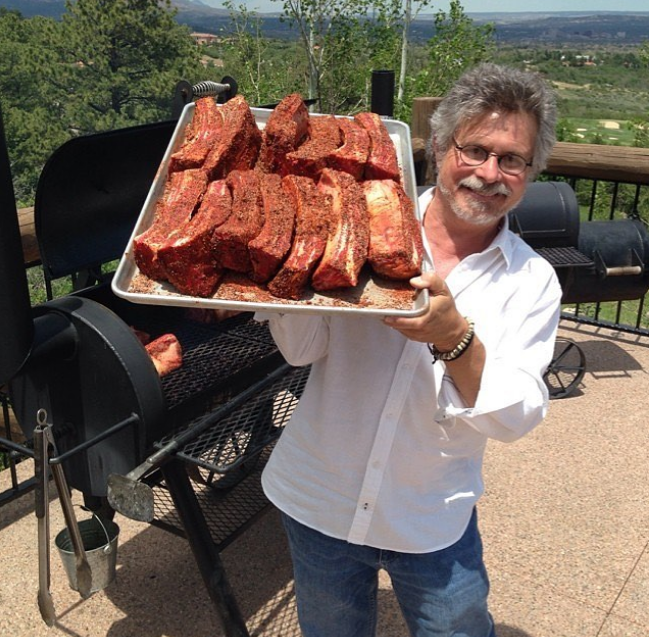 Man in white shirt and glasses holding a large tray of seasoned beef ribs outdoors.