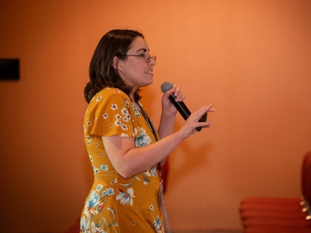 A woman in a yellow floral dress speaks into a handheld microphone in front of an orange wall.