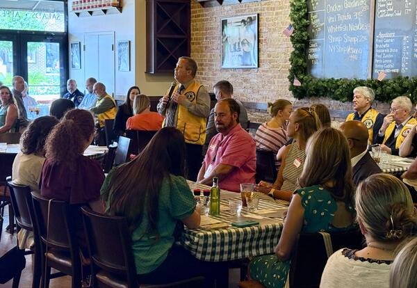Man in yellow Lions Club vest speaks into microphone at a crowded luncheon.
