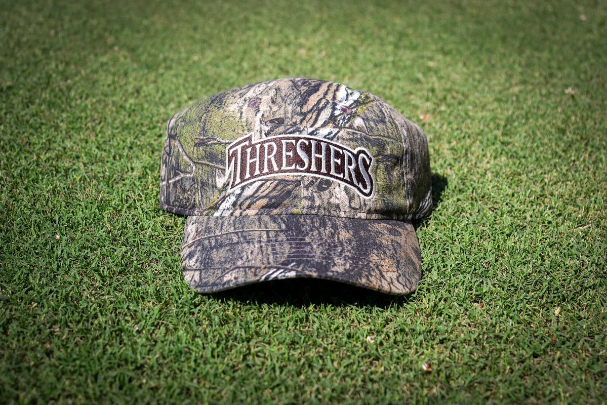 A camouflage baseball cap with 'Threshers' embroidered on the front, resting on green grass.