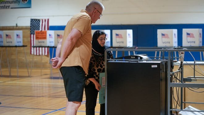 Two people stand at a ballot scanning machine inside a gymnasium polling place with voting booths and American flags.