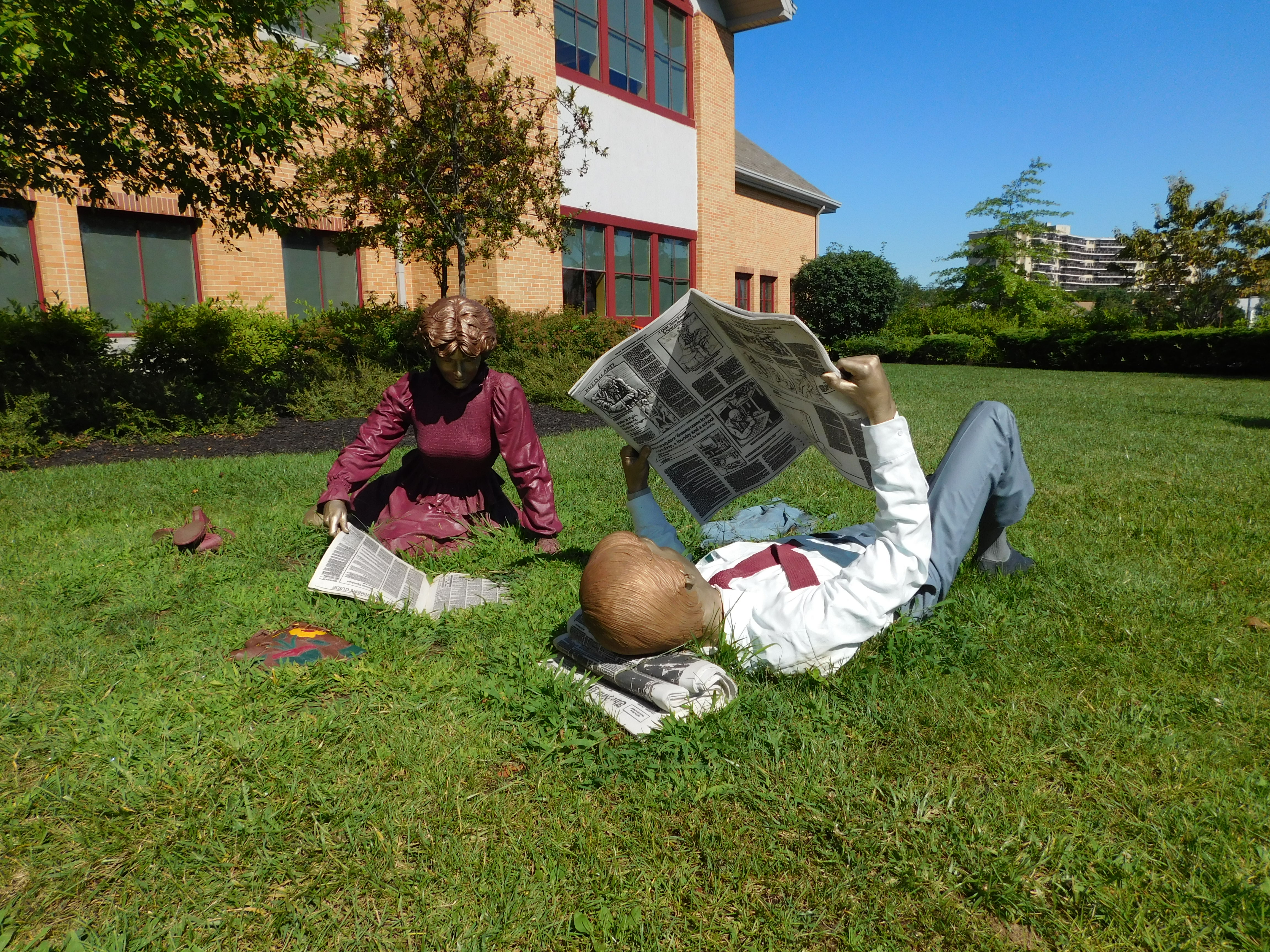 Two bronze sculptures of figures reading newspapers on a grass lawn in front of a brick building.